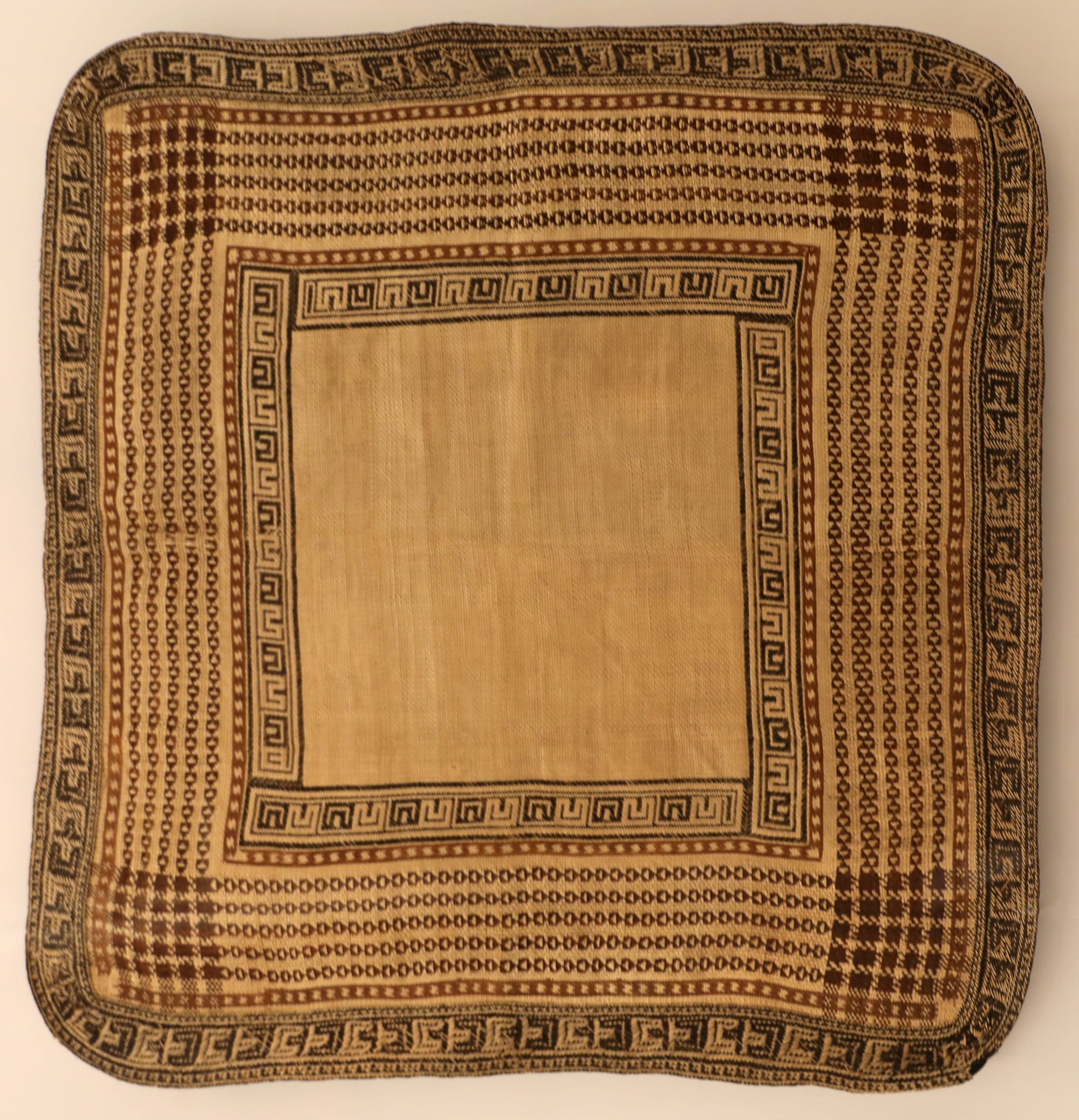 A square woven pandanus mat with a plain pale centre framed by several bands of dark brown geometric patterns, including key-shaped motifs and chequered corners