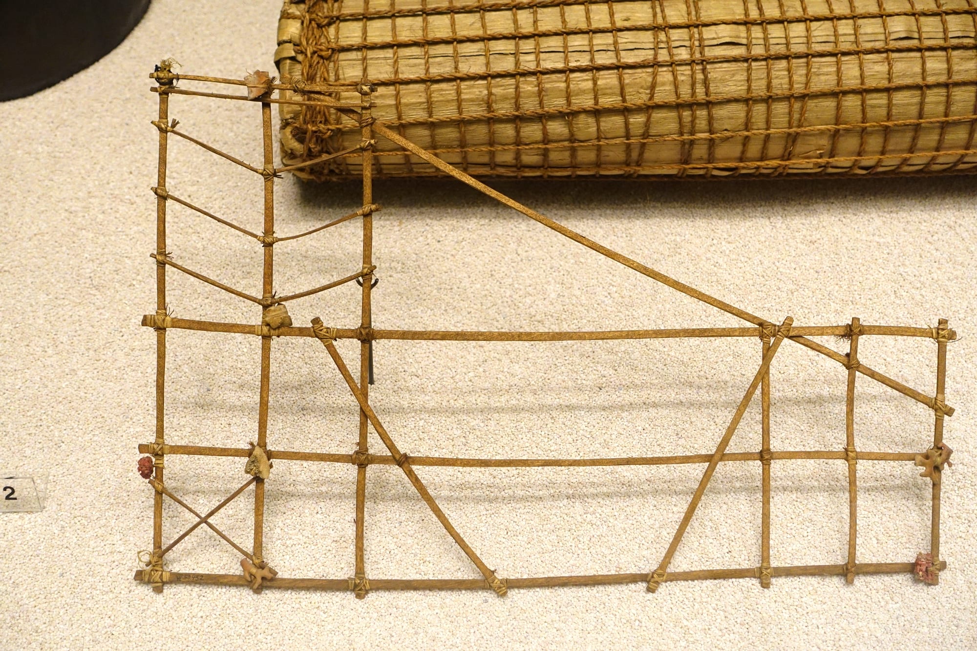 A flat frame of thin lashed sticks forming rectangles and diagonals, with small markers tied at several junctions, displayed in front of a rolled woven mat