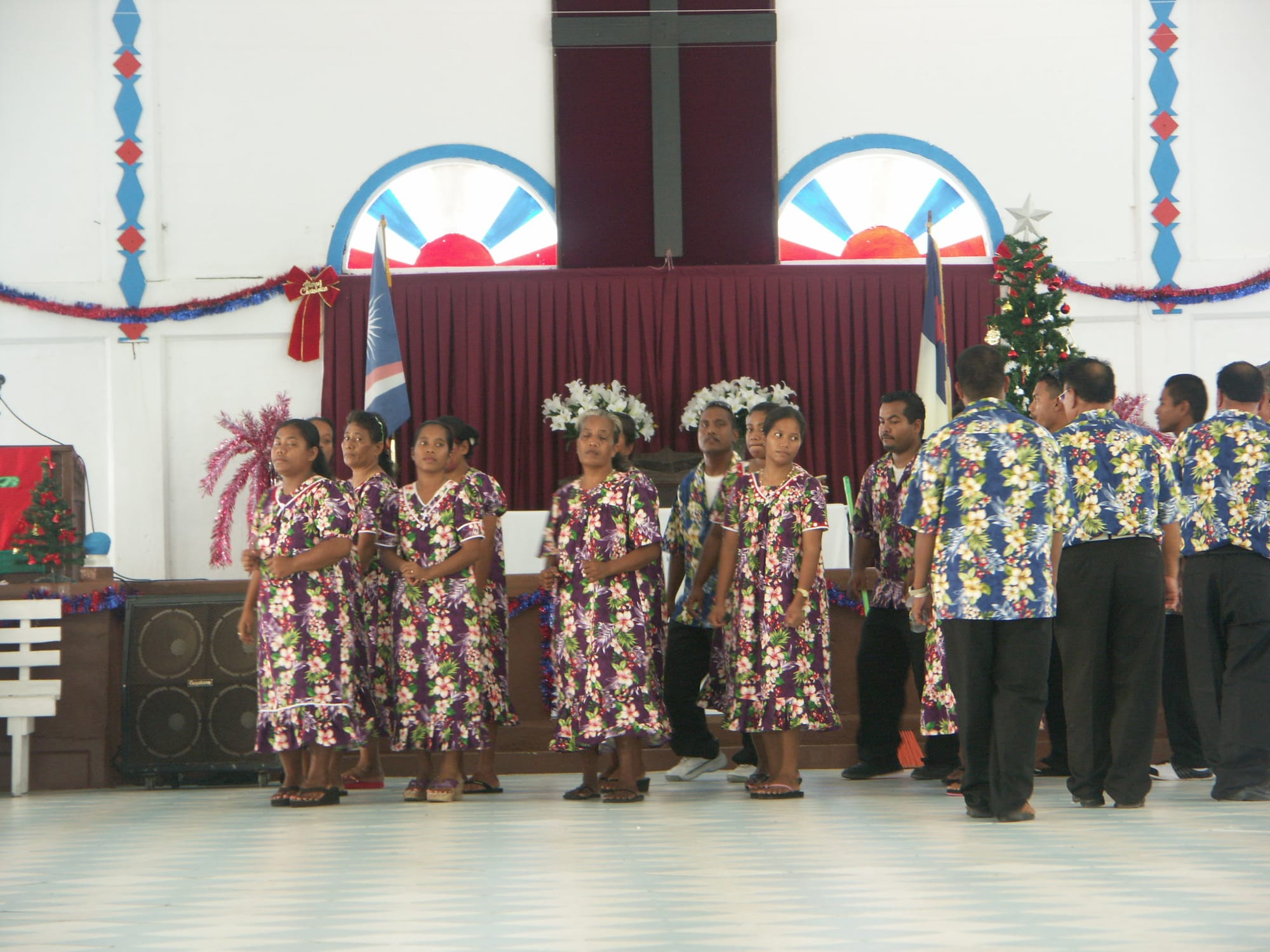 A group of women in matching purple floral dresses and men in matching blue floral shirts stand before a church altar hung with red curtains and a cross, with a Christmas tree and the national flag nearby; two women wear white flower crowns