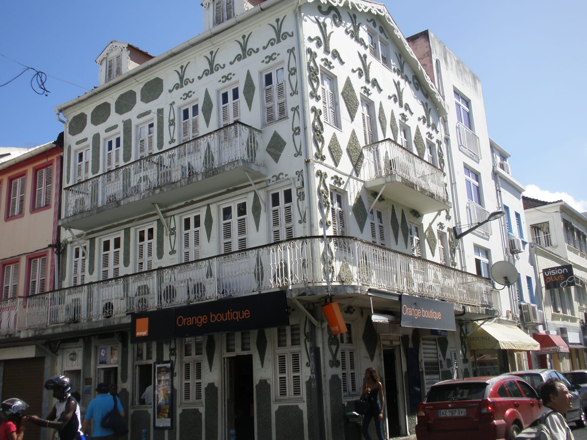 An ornate white corner building with wrought-iron balconies and shuttered windows on a Fort-de-France street, with pedestrians, a motorbike and cars passing below