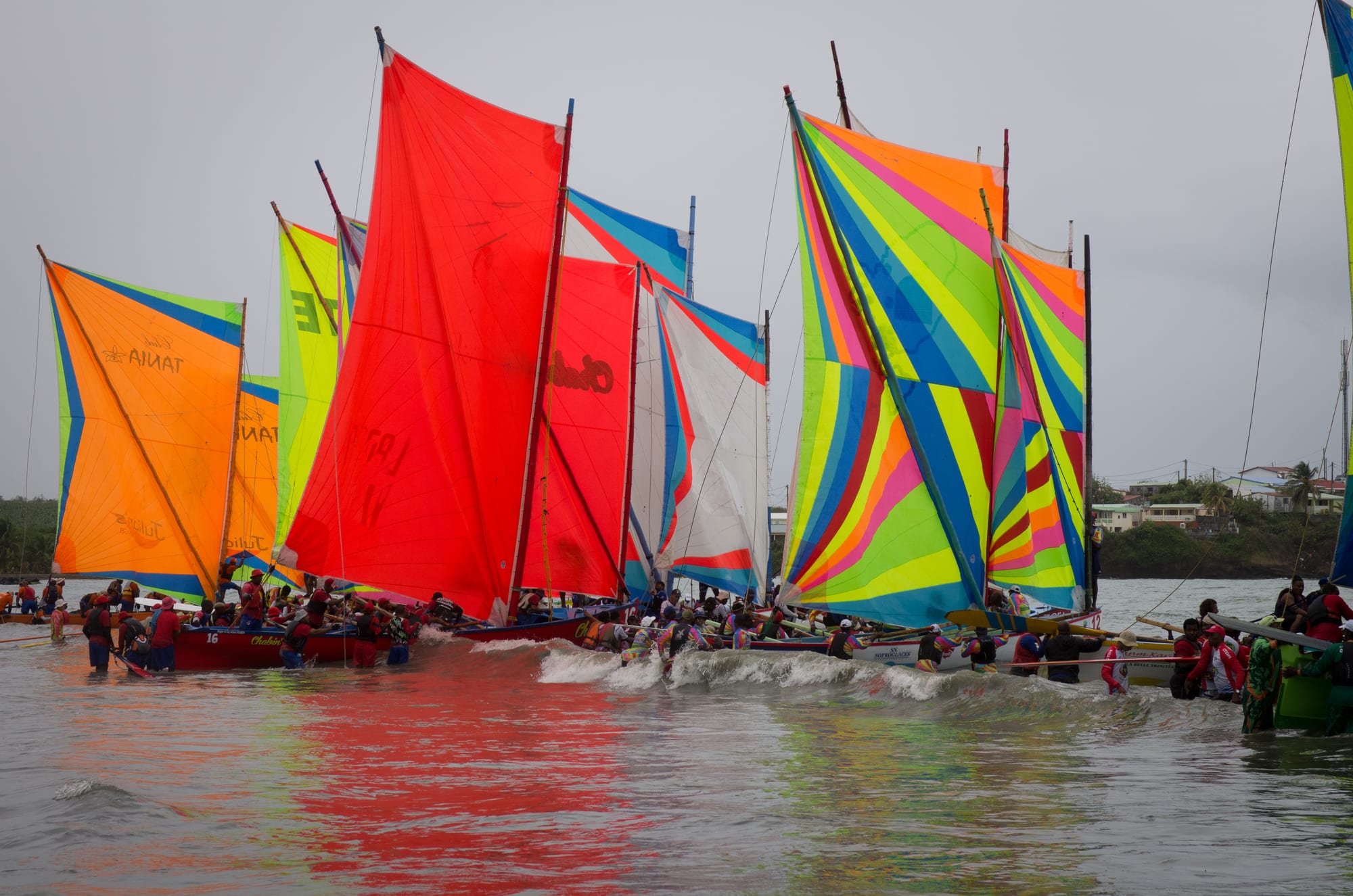 A crowd of traditional sailing boats with tall, brightly coloured sails in red, orange, yellow, blue and green push off from a beach into shallow water, their crews in the waves beside them