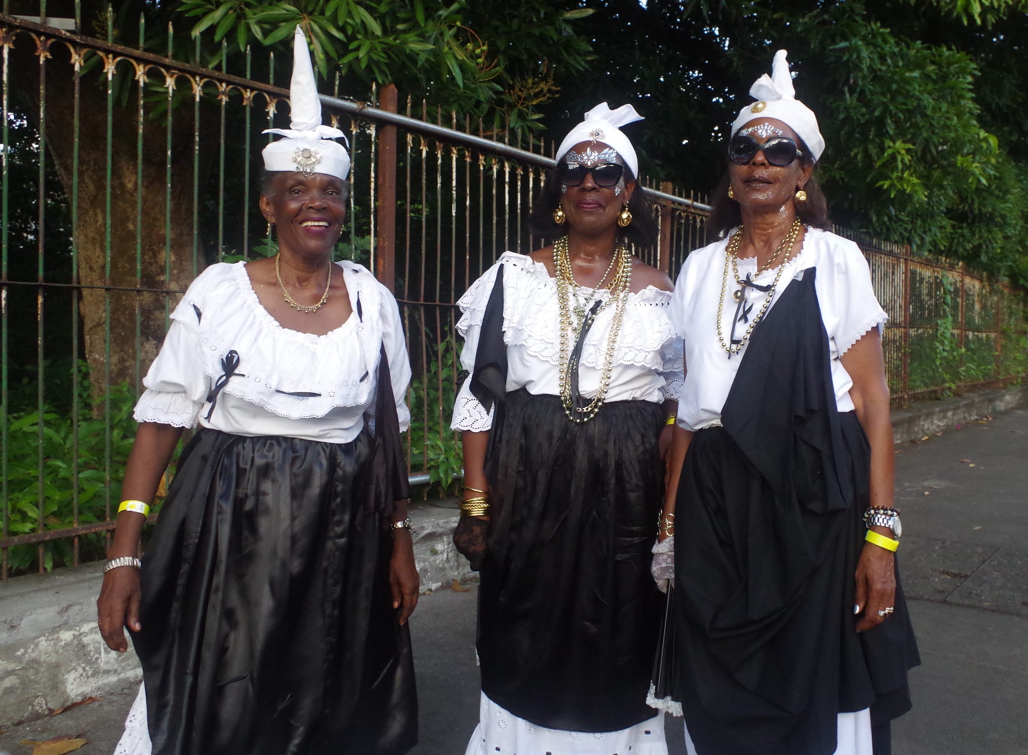 Three smiling women in white lace blouses, long black skirts and black shawls, with tall white head-ties and gold necklaces, stand together on a pavement beside an iron railing