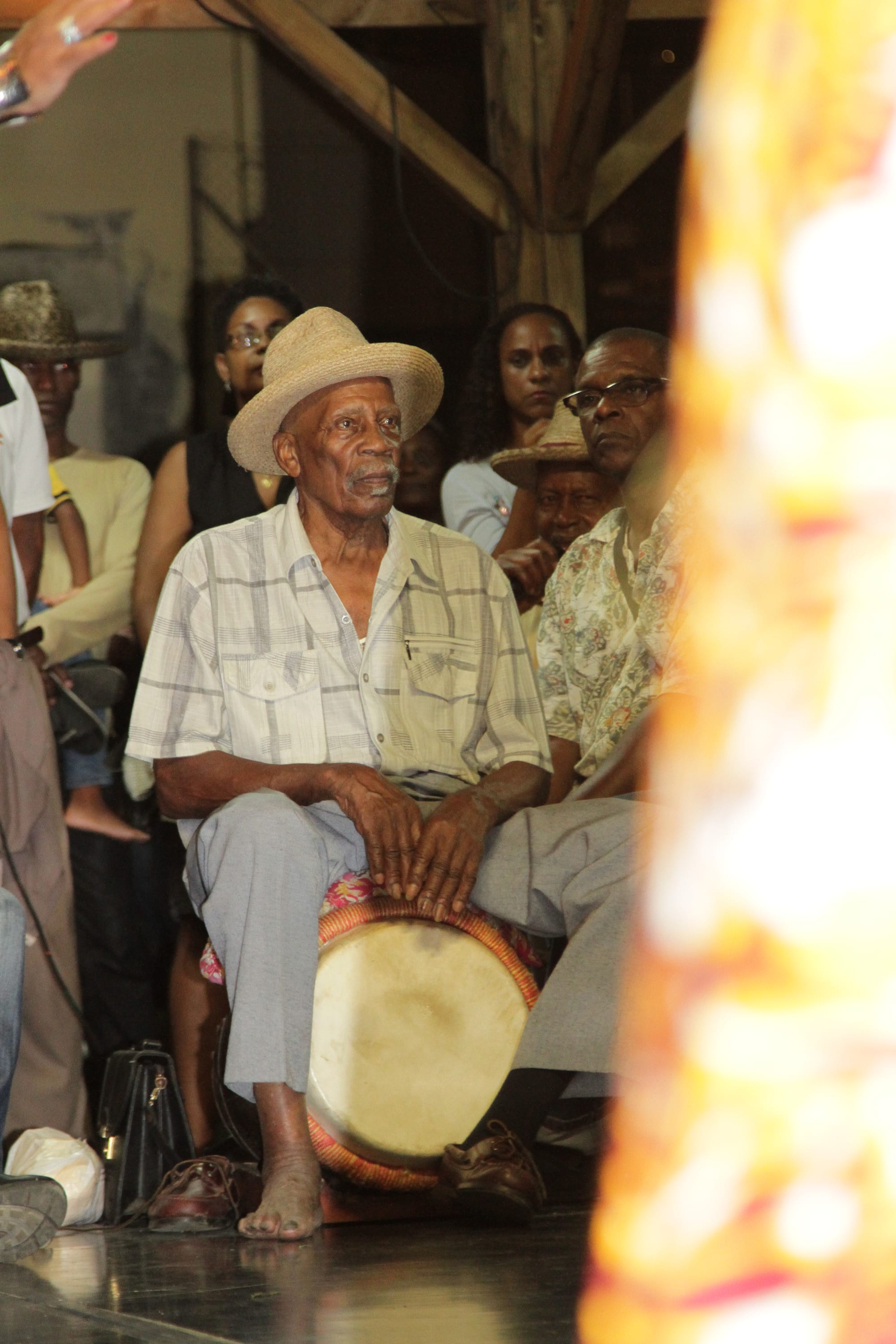 An elderly man in a straw hat and checked short-sleeved shirt sits barefoot with a bèlè drum between his knees, with an audience seated behind him