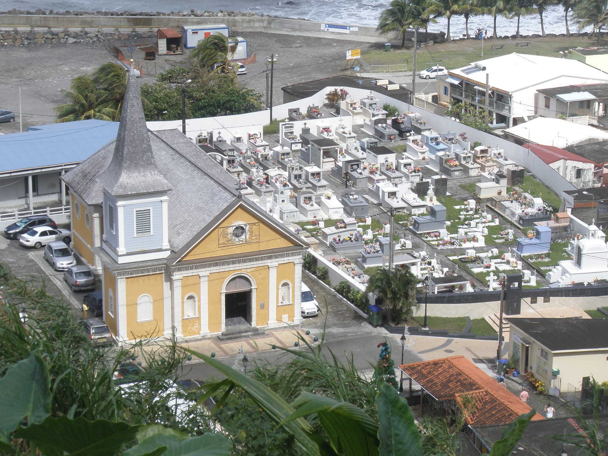 A yellow church with a grey slate roof and pointed bell tower beside a hillside cemetery crowded with white-painted tombs decorated with flowers, with the sea beyond