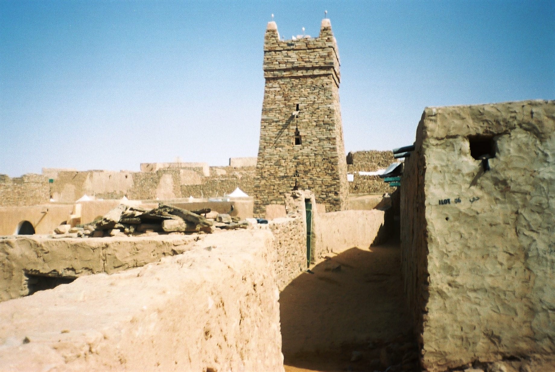 A mud-and-stone mosque with a square minaret rising from a desert town partly buried in sand