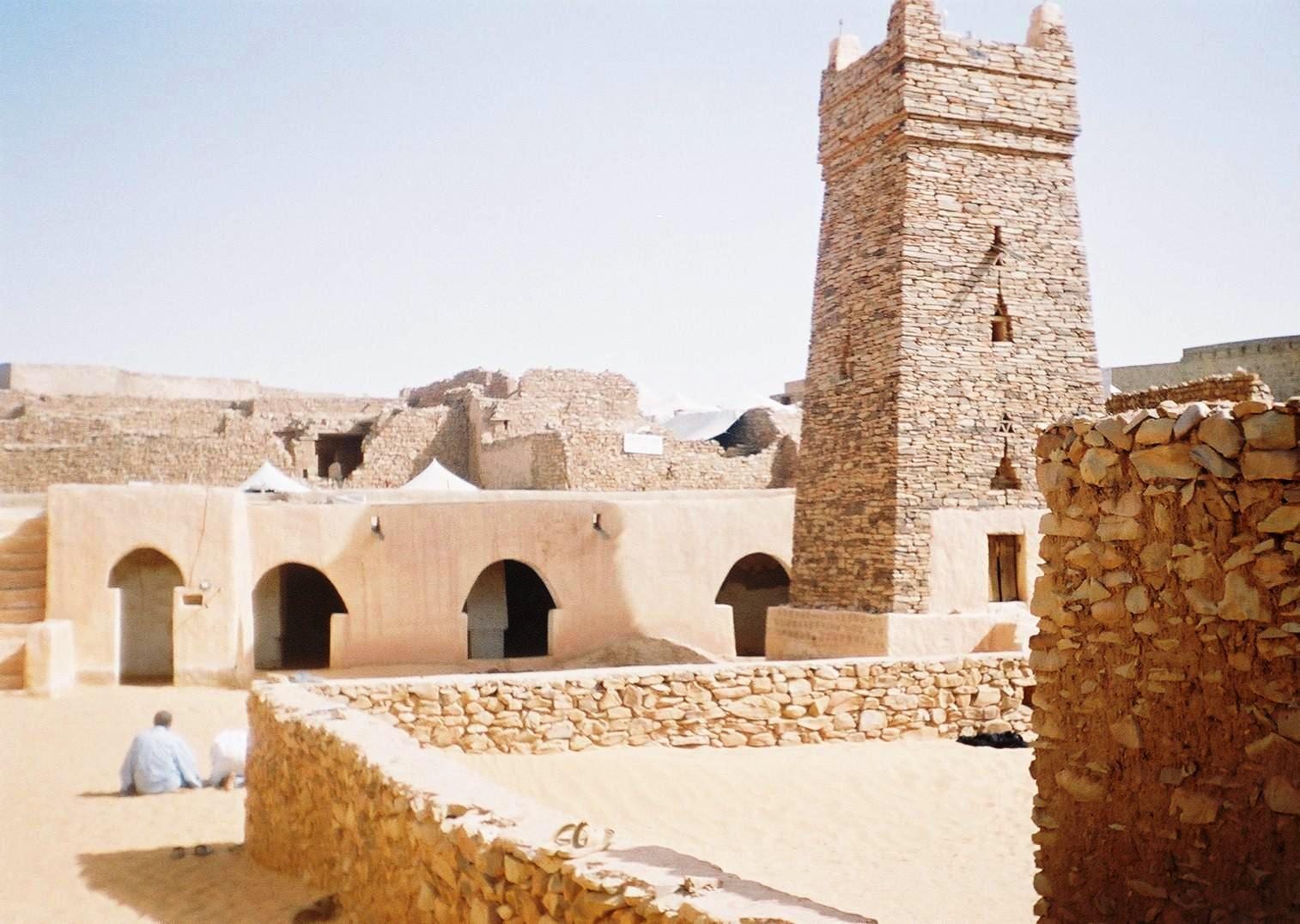 A weathered mud mosque with a square tower, its base partly covered by drifting sand