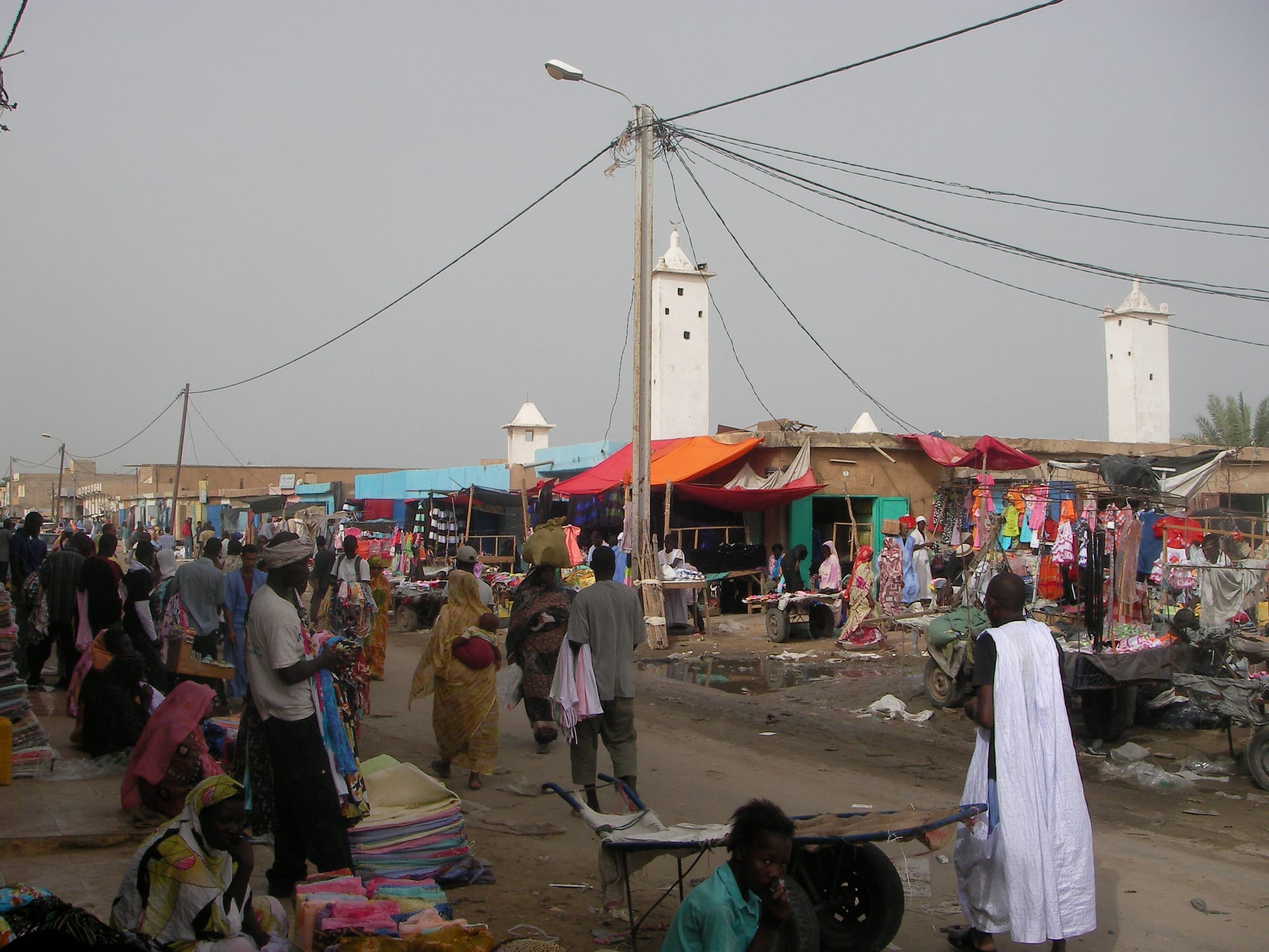 A crowded street market in Nouakchott with vendor stalls, coloured awnings and two mosque minarets in the background