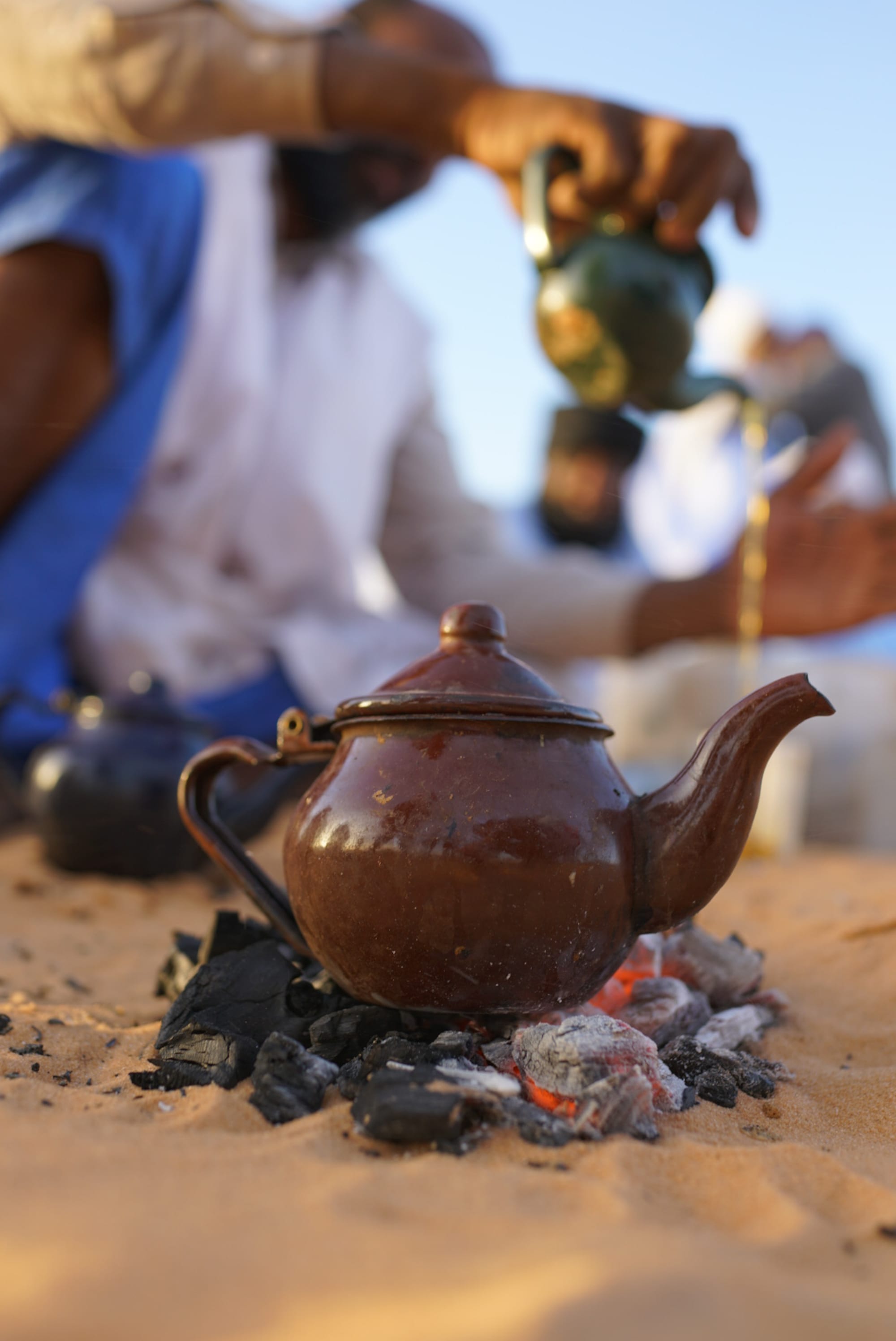 A small teapot brewing on hot coals in the sand, with a man pouring tea from another kettle in the background