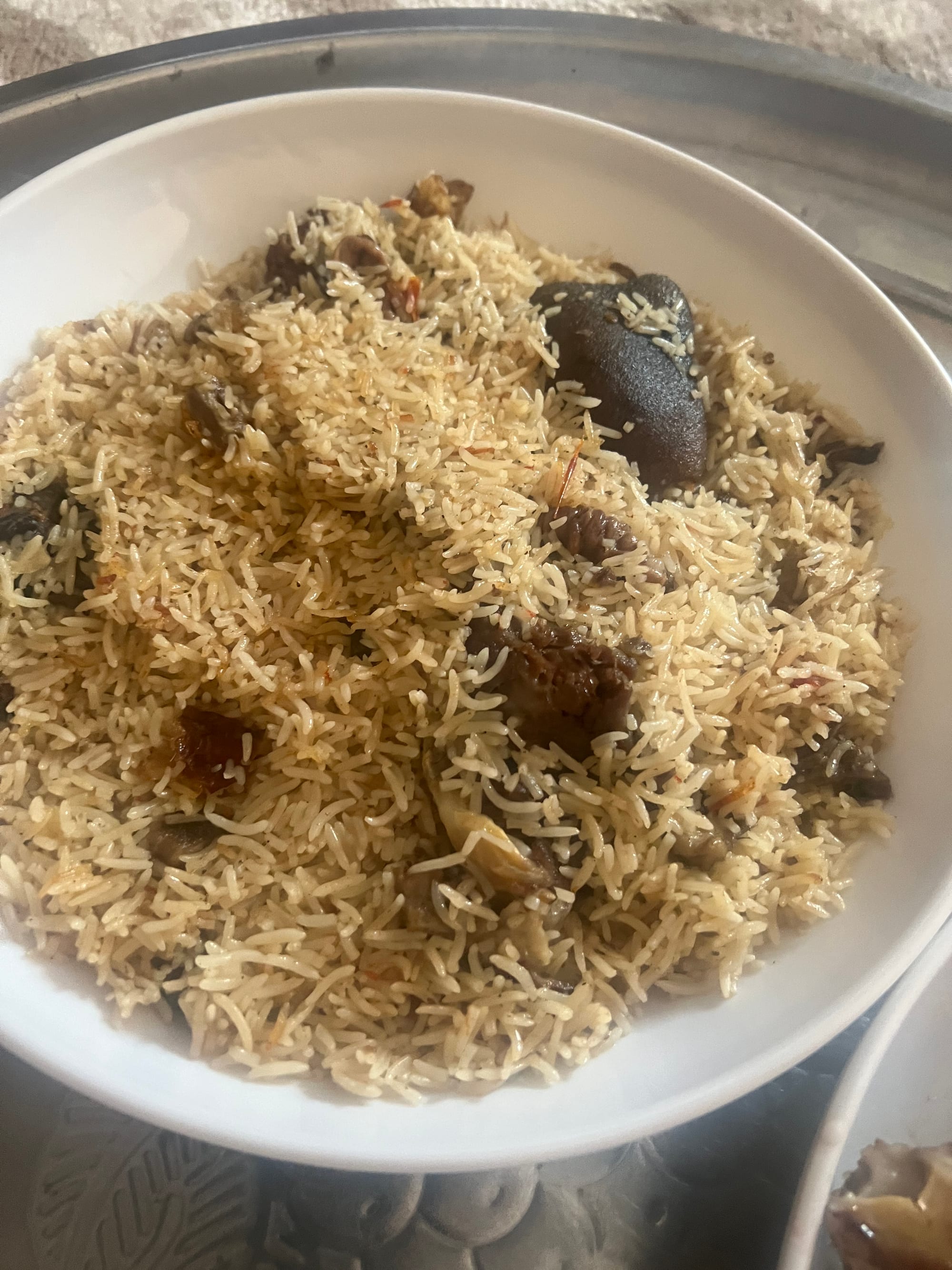 A large white bowl heaped with golden rice and pieces of dark cooked meat, resting on a round metal tray