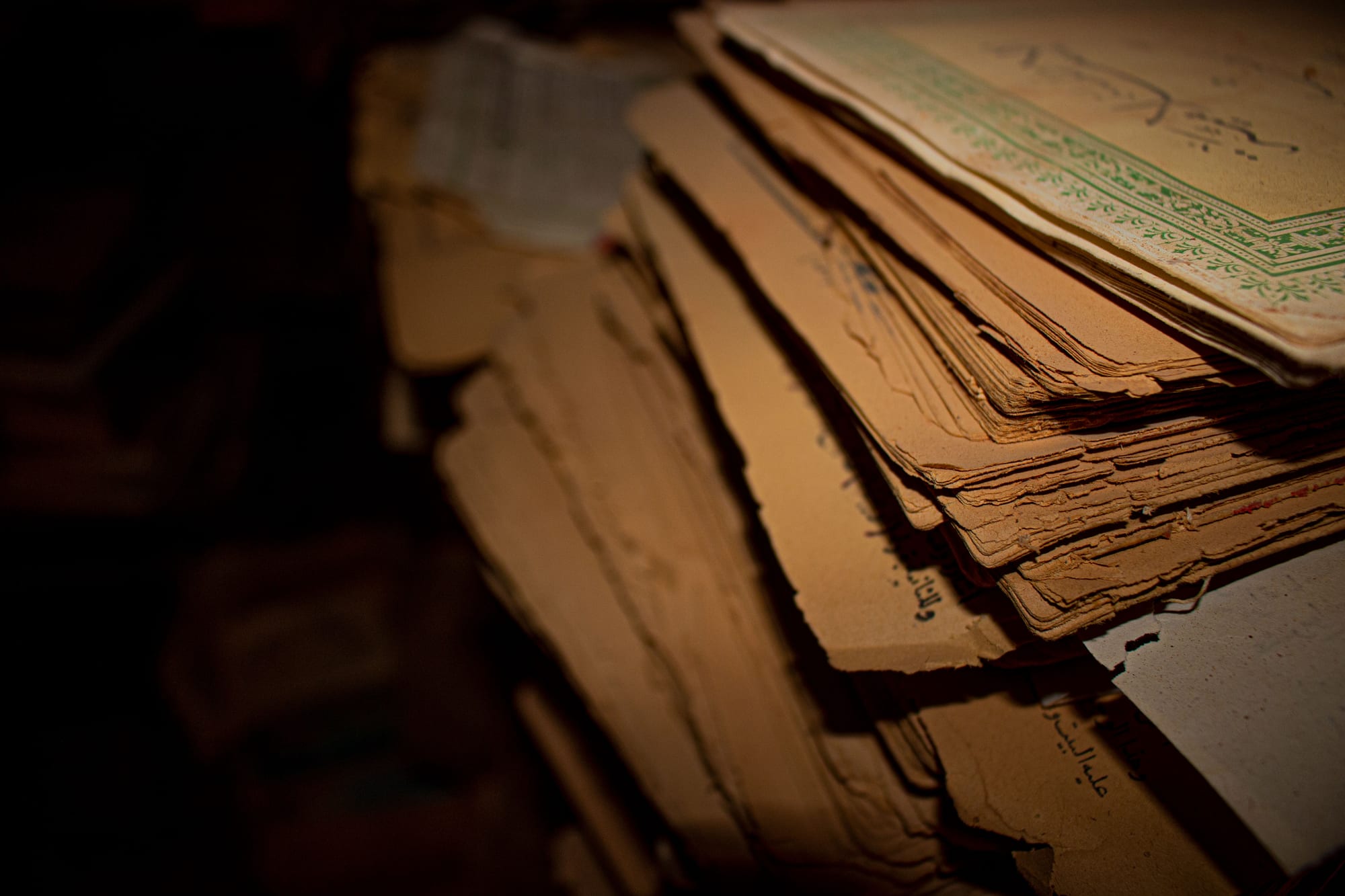 A close view of a stack of old manuscript pages with frayed, yellowed edges and handwritten Arabic script, one decorated with a green border, lit softly against a dark background