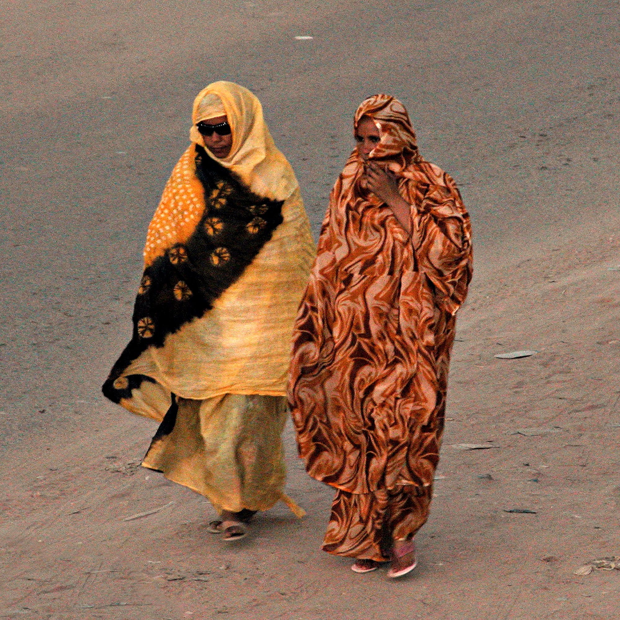 Two women walk along a sandy road wrapped in long malahfa, one in yellow and black with a sunburst pattern, the other in swirling brown and orange, with the cloth drawn over their heads