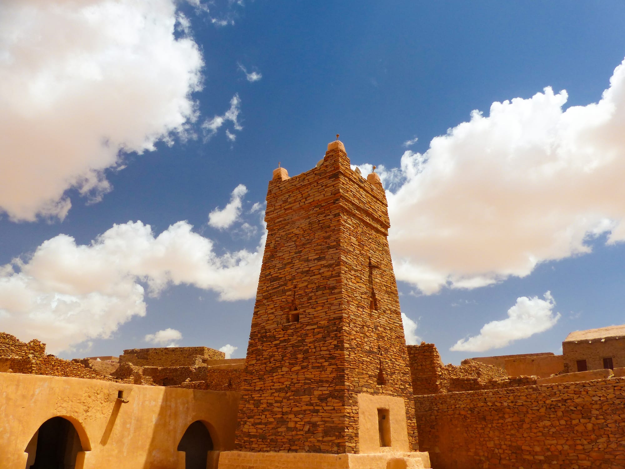A square minaret of layered flat stones, topped with small pointed finials, rises above ochre walls and a courtyard with arched doorways under a blue sky with white clouds