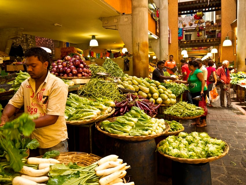 A vendor arranges fresh vegetables at a covered market stall in Port Louis, with shoppers browsing baskets of produce behind him