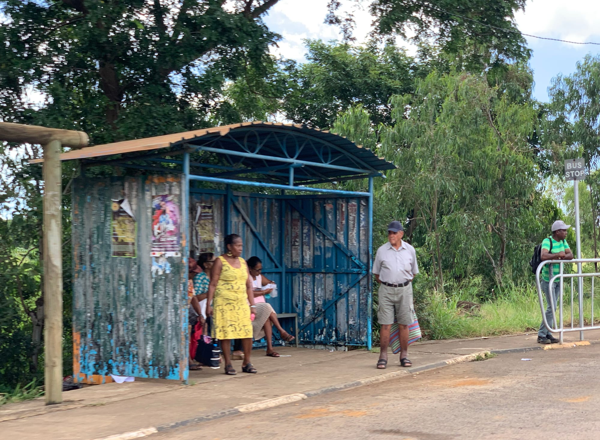 People waiting at a simple roadside bus shelter beside a tree-lined road in Bambous, Mauritius