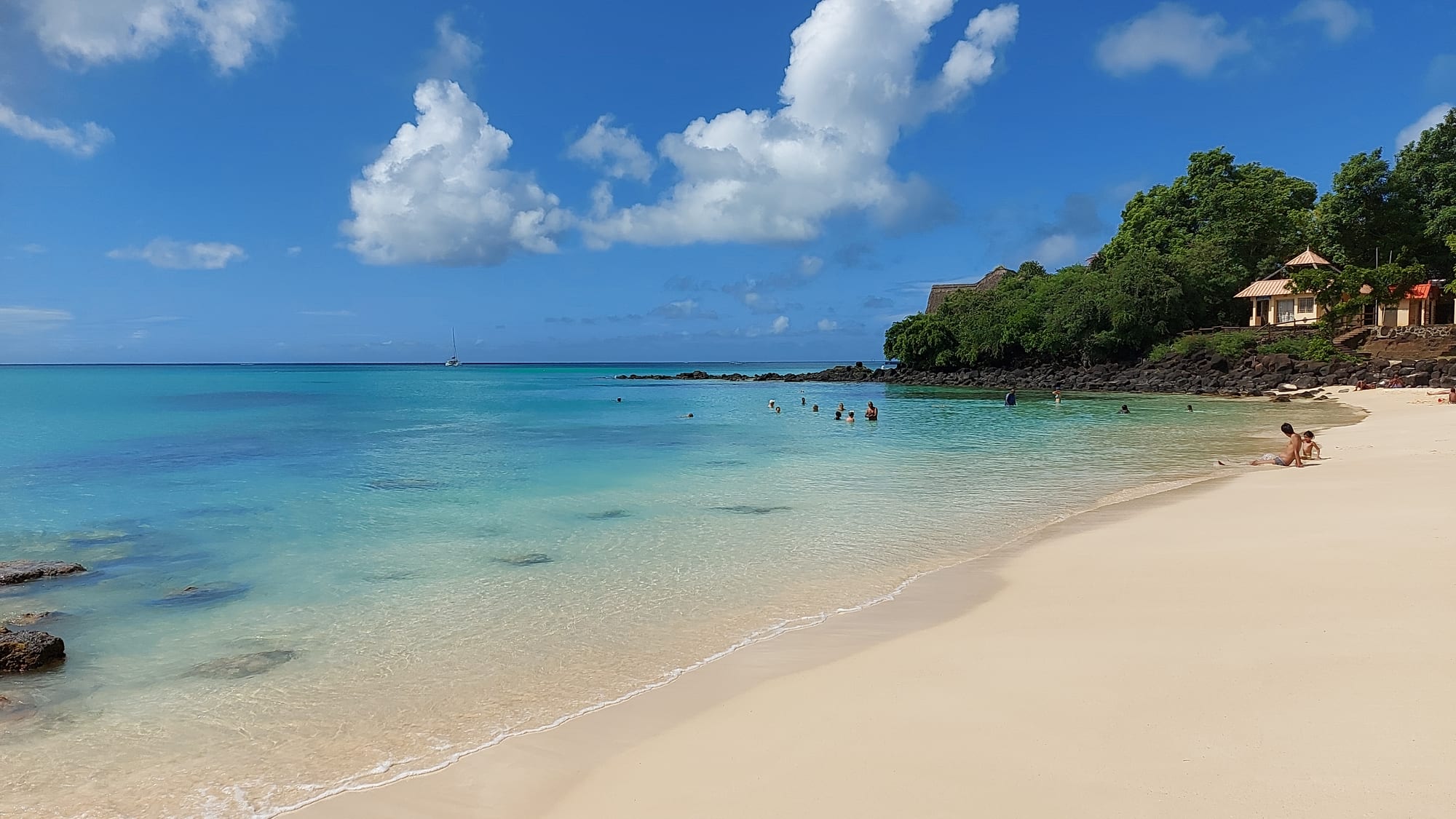 People swimming and sitting at La Cuvette, a public white-sand beach at Grand Baie, with clear turquoise water and palm trees