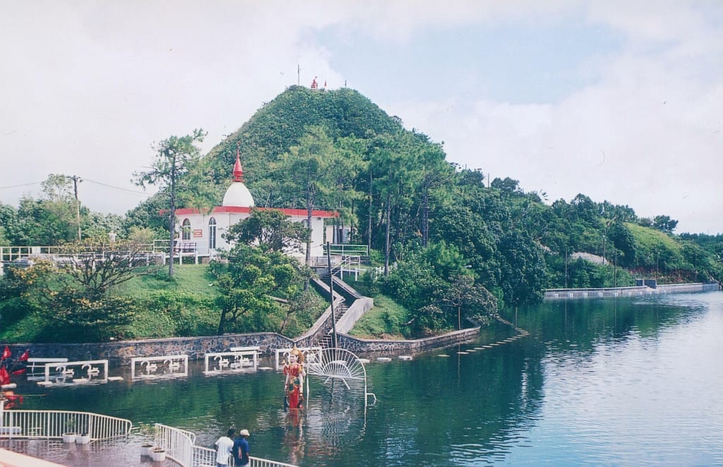 A brightly painted Hindu temple complex beside a crater lake, with a large statue nearby