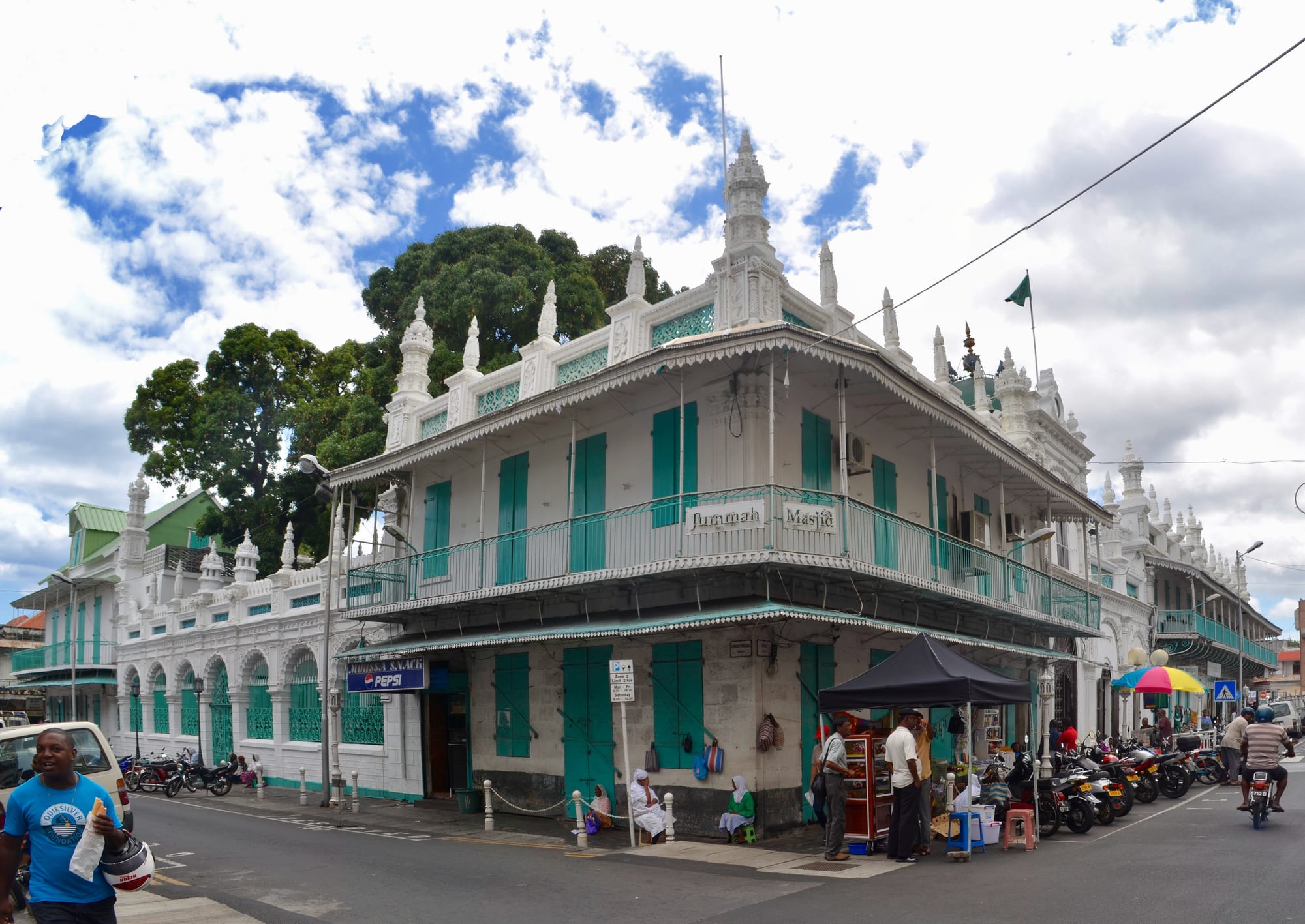 A white two-storey mosque building with ornate pinnacles, green shutters and a long balcony on a street corner, with parked motorbikes and people on the pavement
