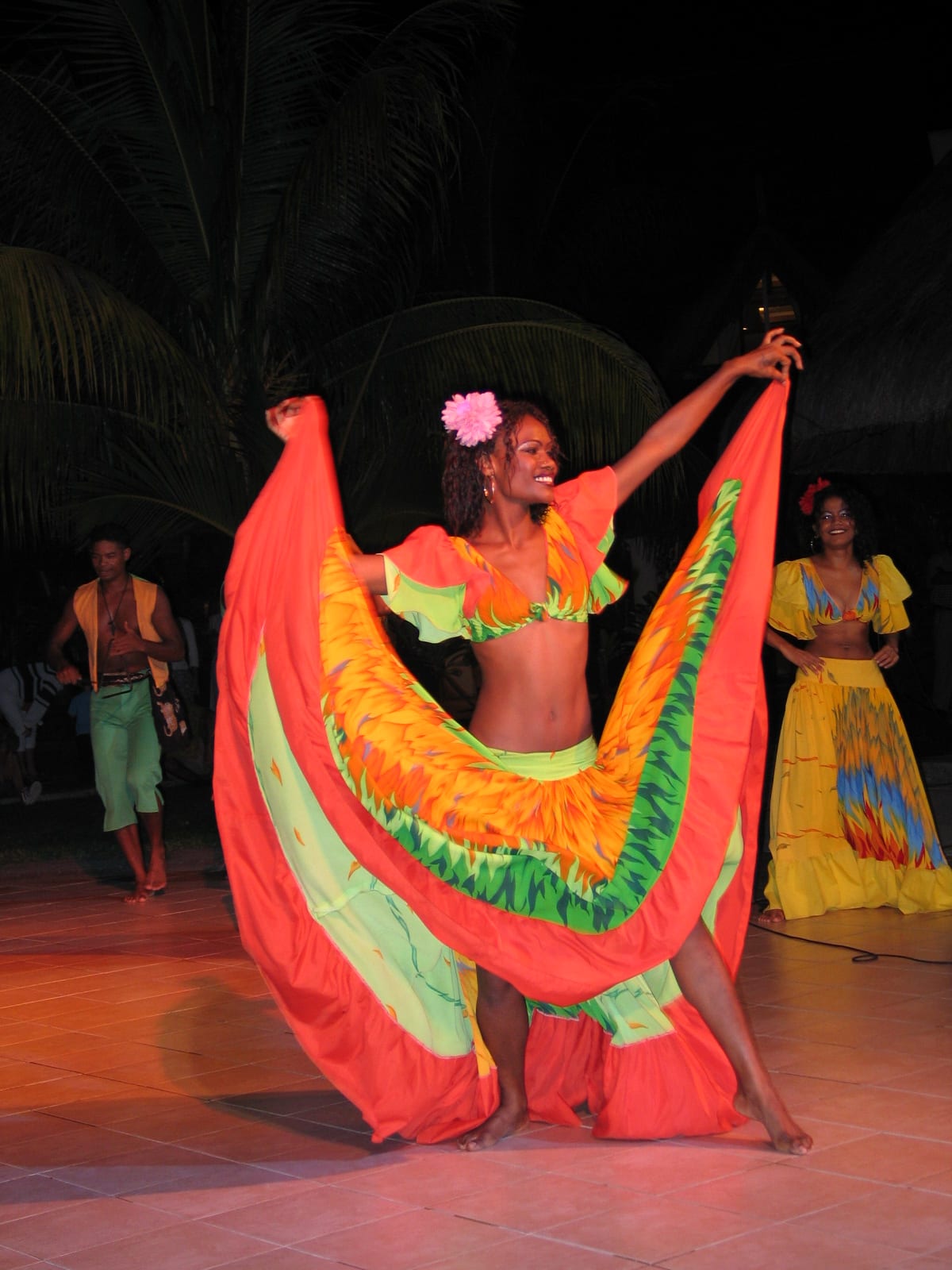 A dancer with a pink flower in her hair holds out the edges of a wide orange, green and red skirt, with another dancer in yellow and a male dancer behind her under palm trees at night