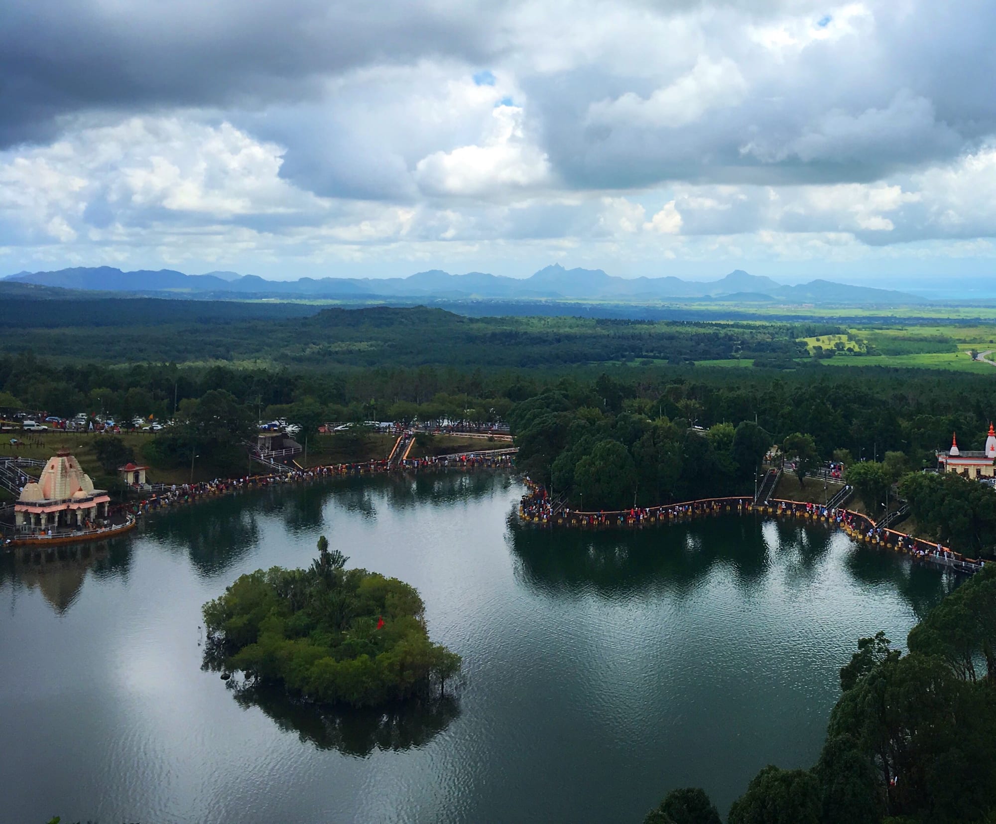 A crater lake seen from above, with a small wooded island in the middle, a pale temple on the left shore and long lines of people along the lakeside walkways, with forest, plains and mountains beyond