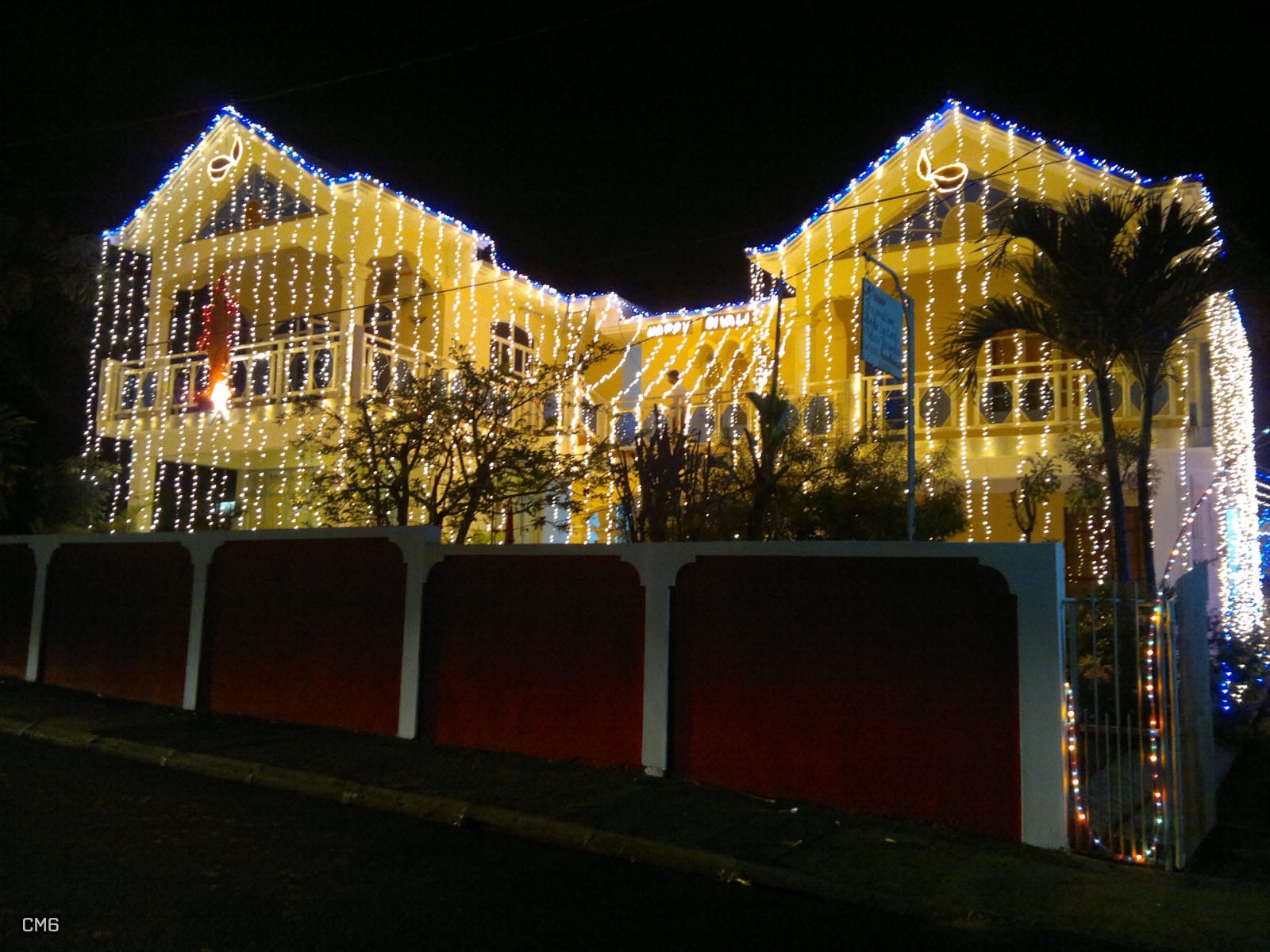 A two-storey yellow house at night covered in hanging strands of white lights, with lamp shapes outlined in lights on the gables and a red and white wall in front