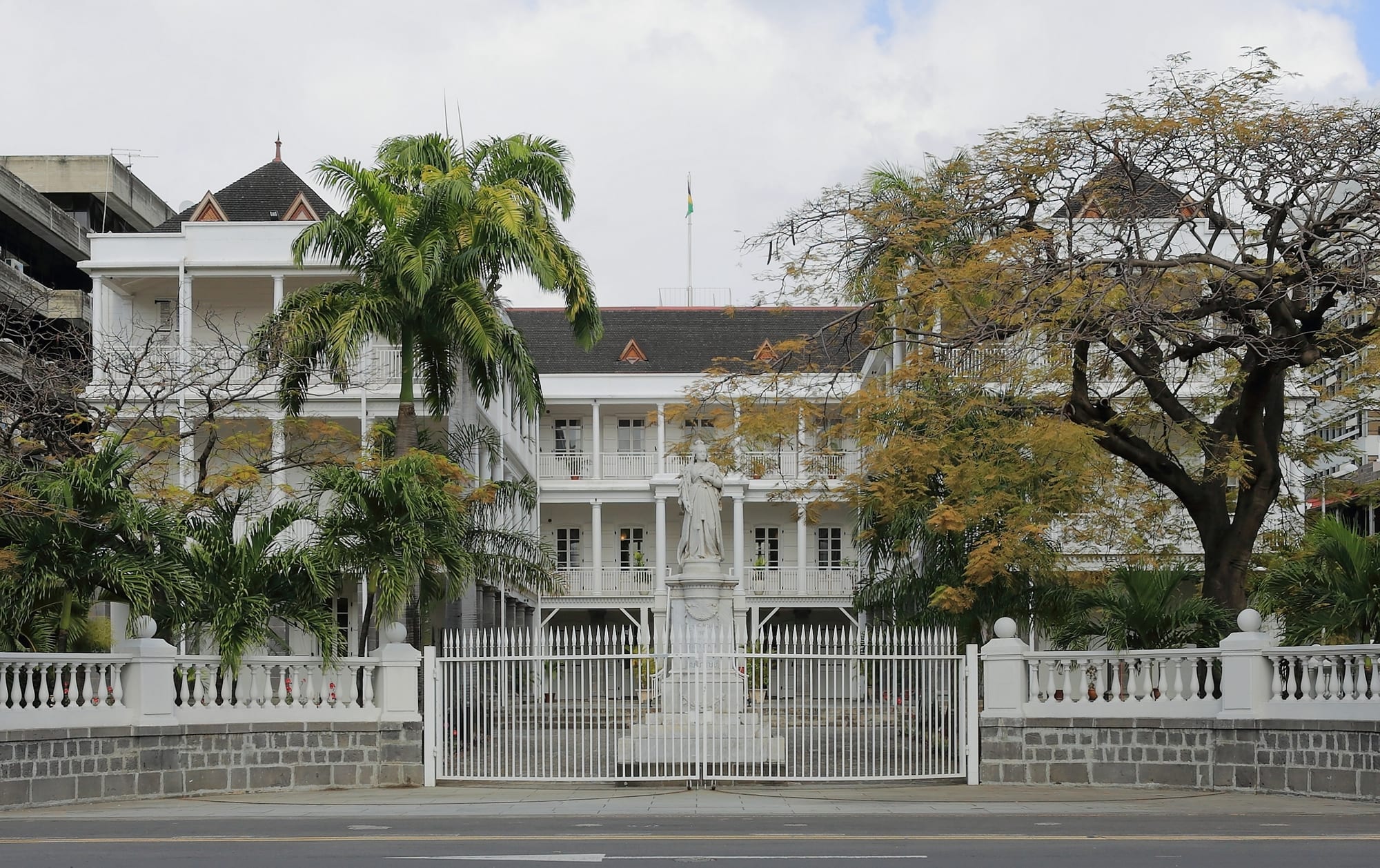 A white colonial building with tiers of verandas behind a white gate, a statue on a plinth in its forecourt and palm trees on either side