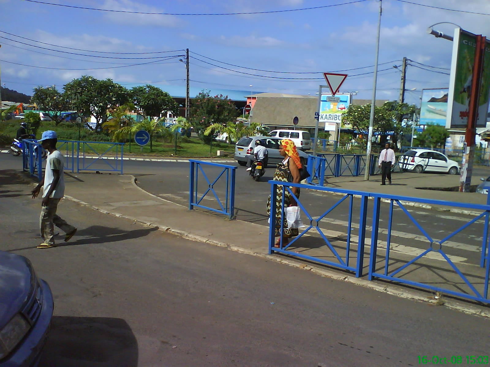 A street junction in Mamoudzou with blue pedestrian railings, people crossing on foot, and cars and a motorbike passing through