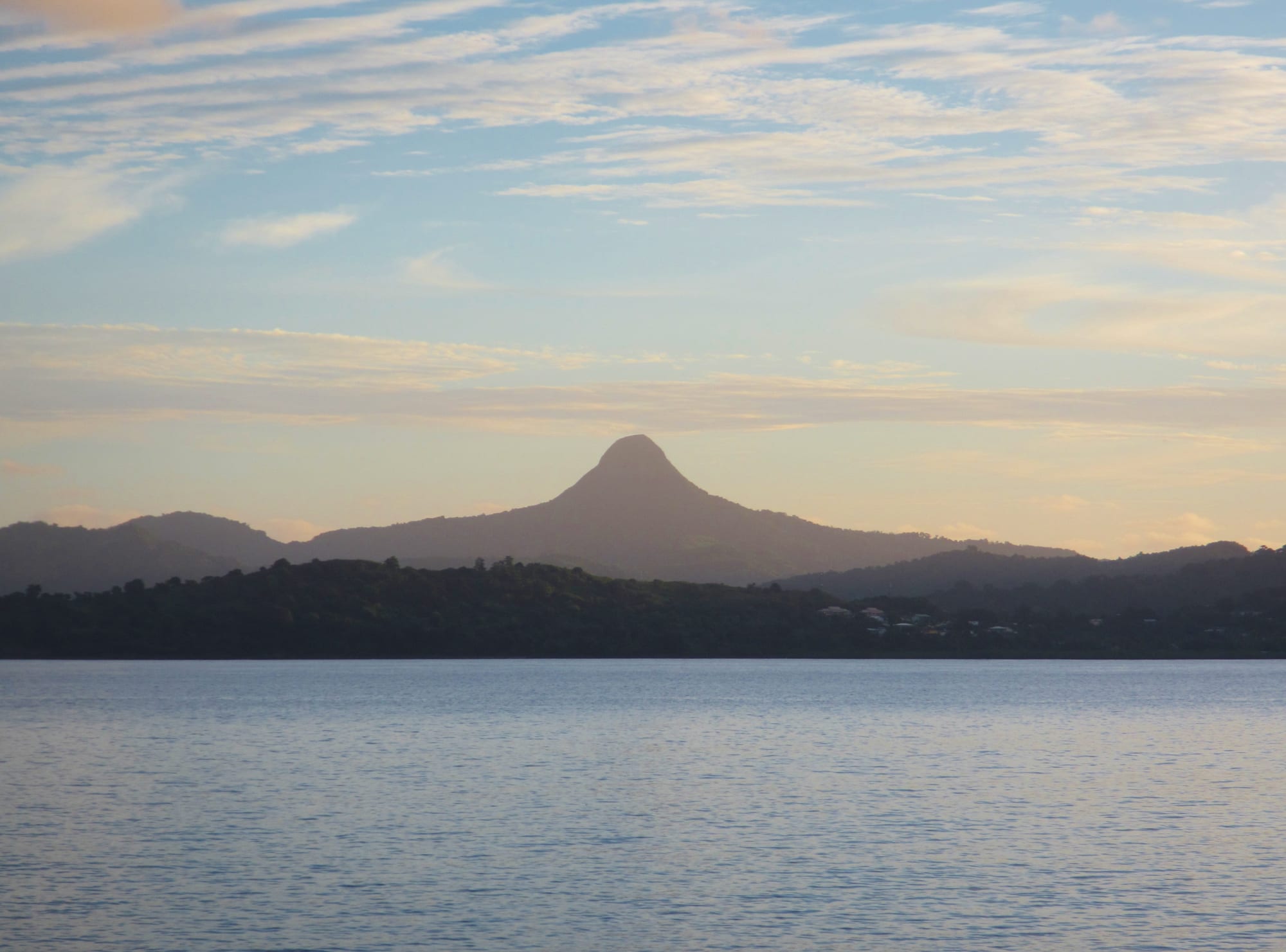 A steep, pointed volcanic peak covered in forest rising above lower green hills
