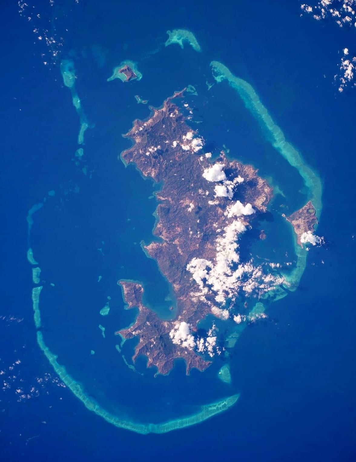 A satellite view of an island fully encircled by a pale turquoise barrier reef