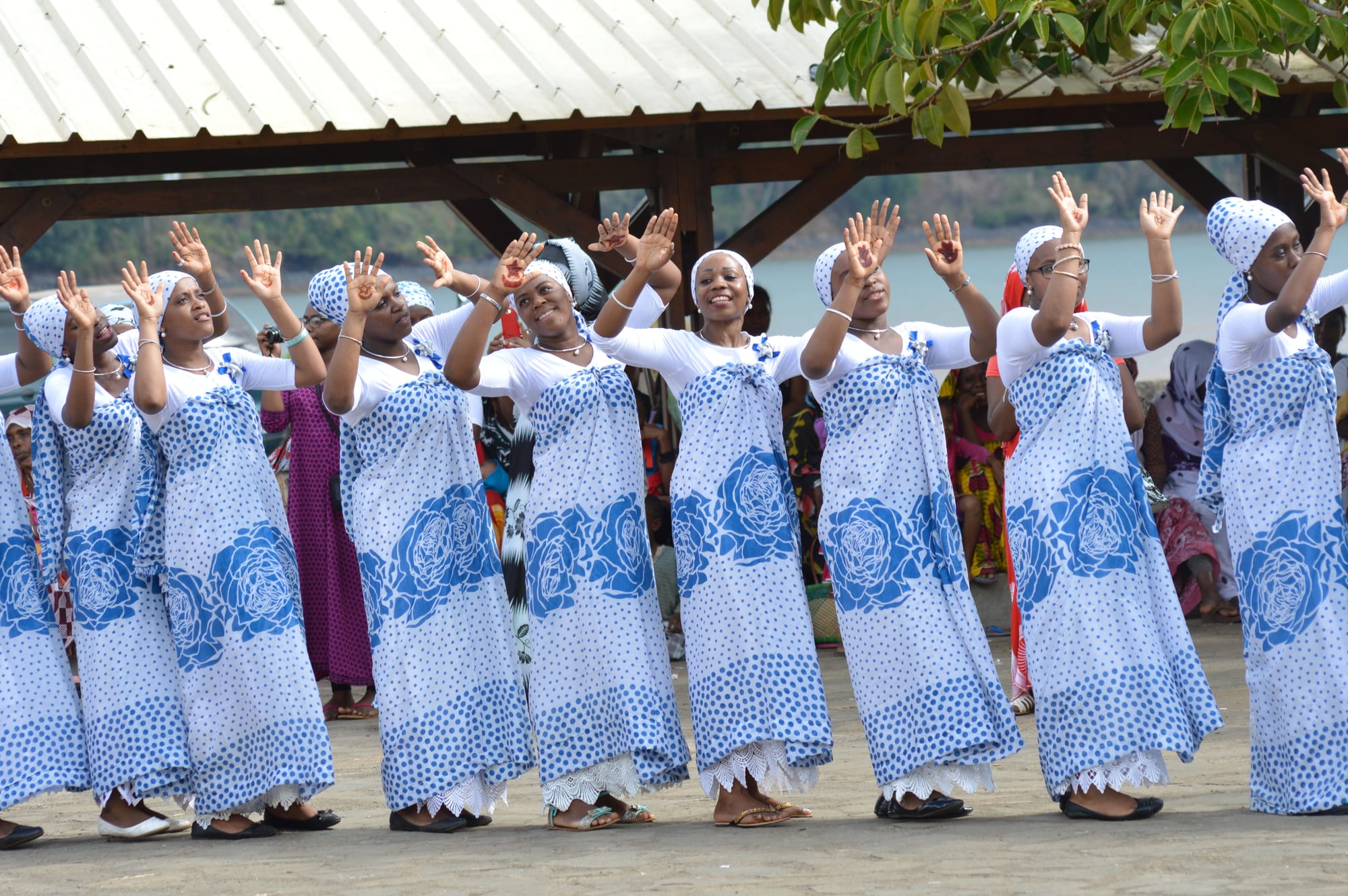A line of women in matching white tops, blue-and-white patterned wraps and headscarves raise their hands, palms outwards, under an open-sided shelter