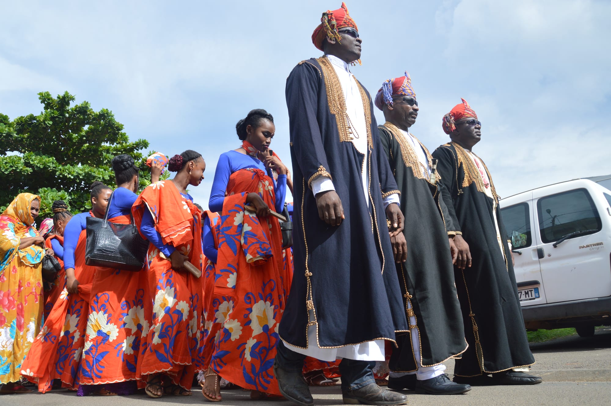 Three men in long dark robes with gold embroidery and red and gold turbans walk along a street beside a line of young women in matching orange and blue wraps with floral patterns