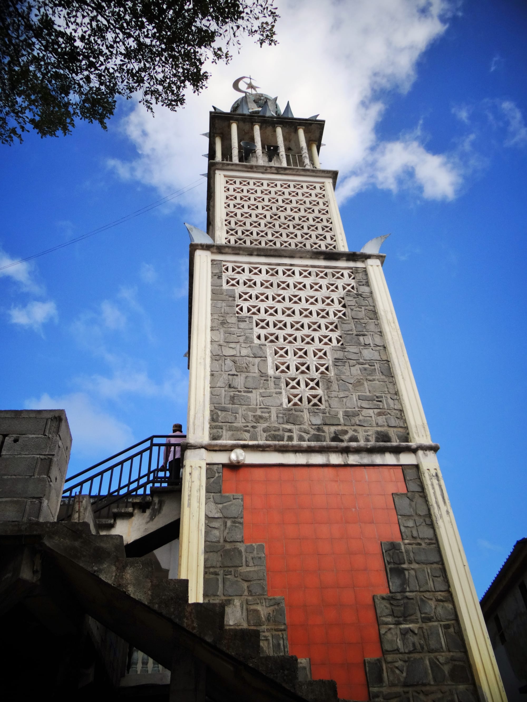 A tall square minaret of grey stone and white trim, with a panel of orange tiles at its base, pierced decorative blockwork above and a crescent at the top, seen from below against a blue sky