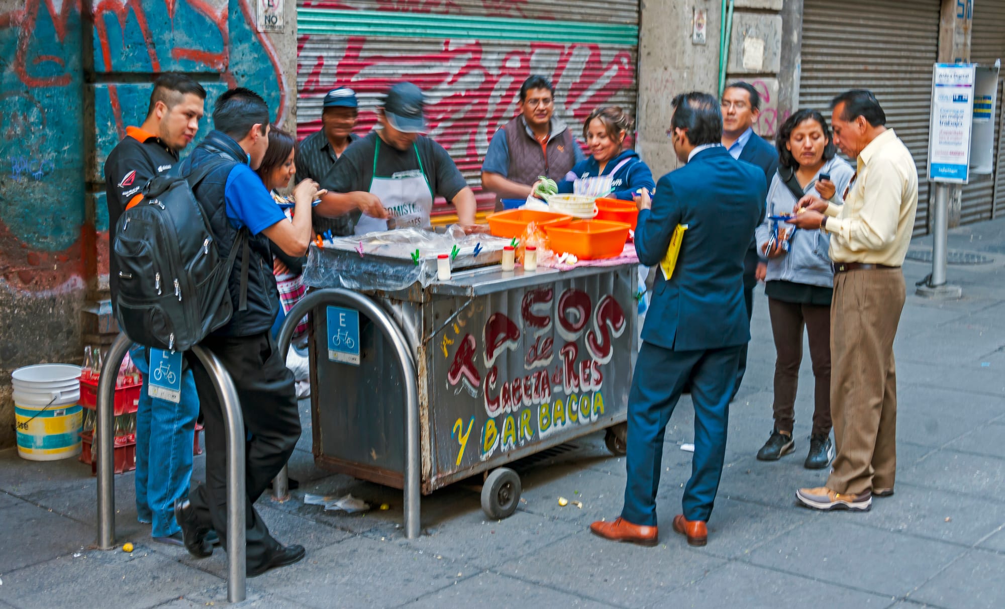 A group of people eating and buying tacos from a metal street cart painted with the words Tacos de Cabeza de Res y Barbacoa