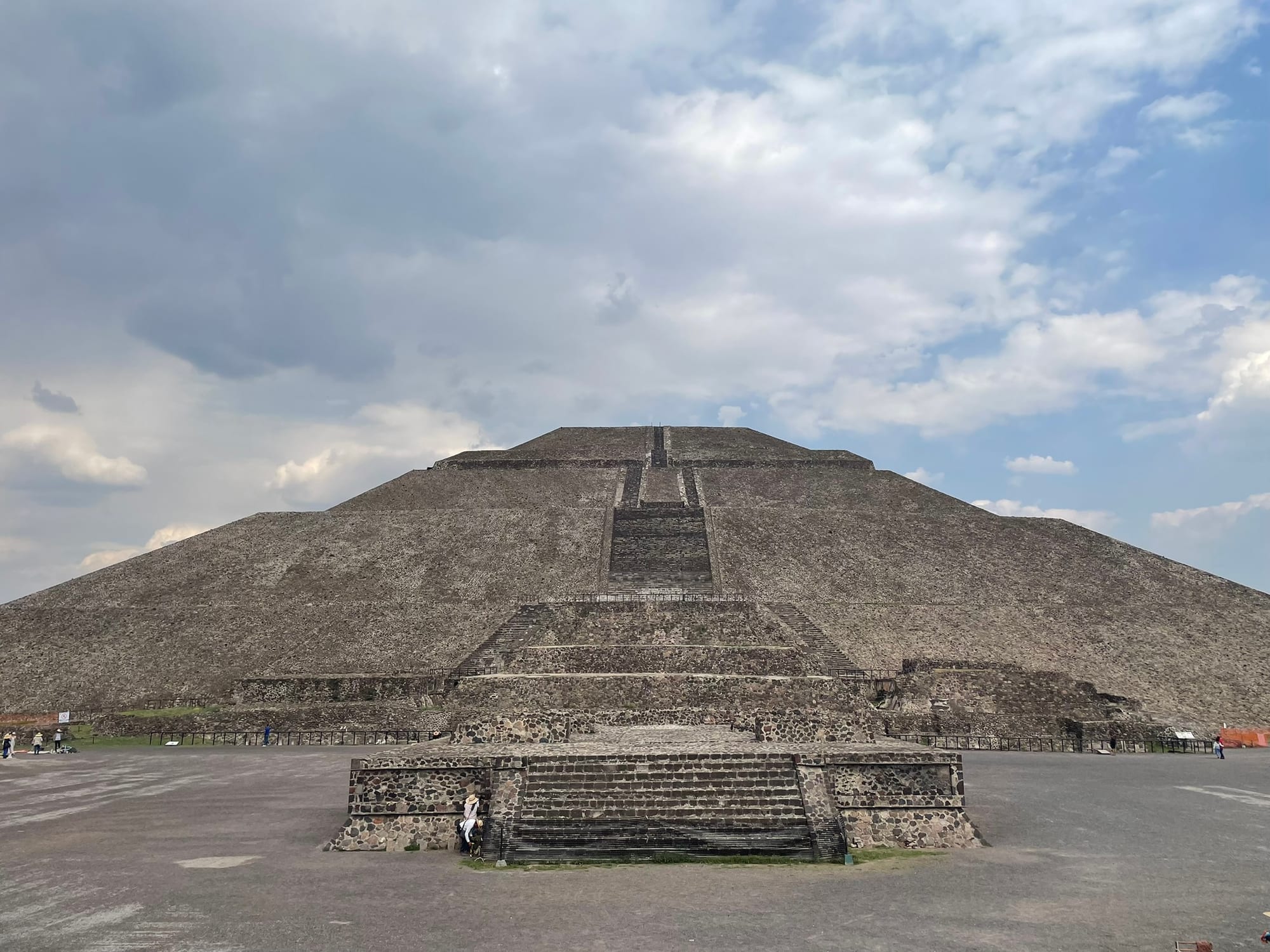 A massive stepped pyramid rising above a stone plaza, clouds behind