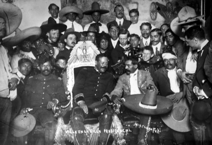 Pancho Villa seated in an ornate chair with Emiliano Zapata beside him, surrounded by armed followers, 1914