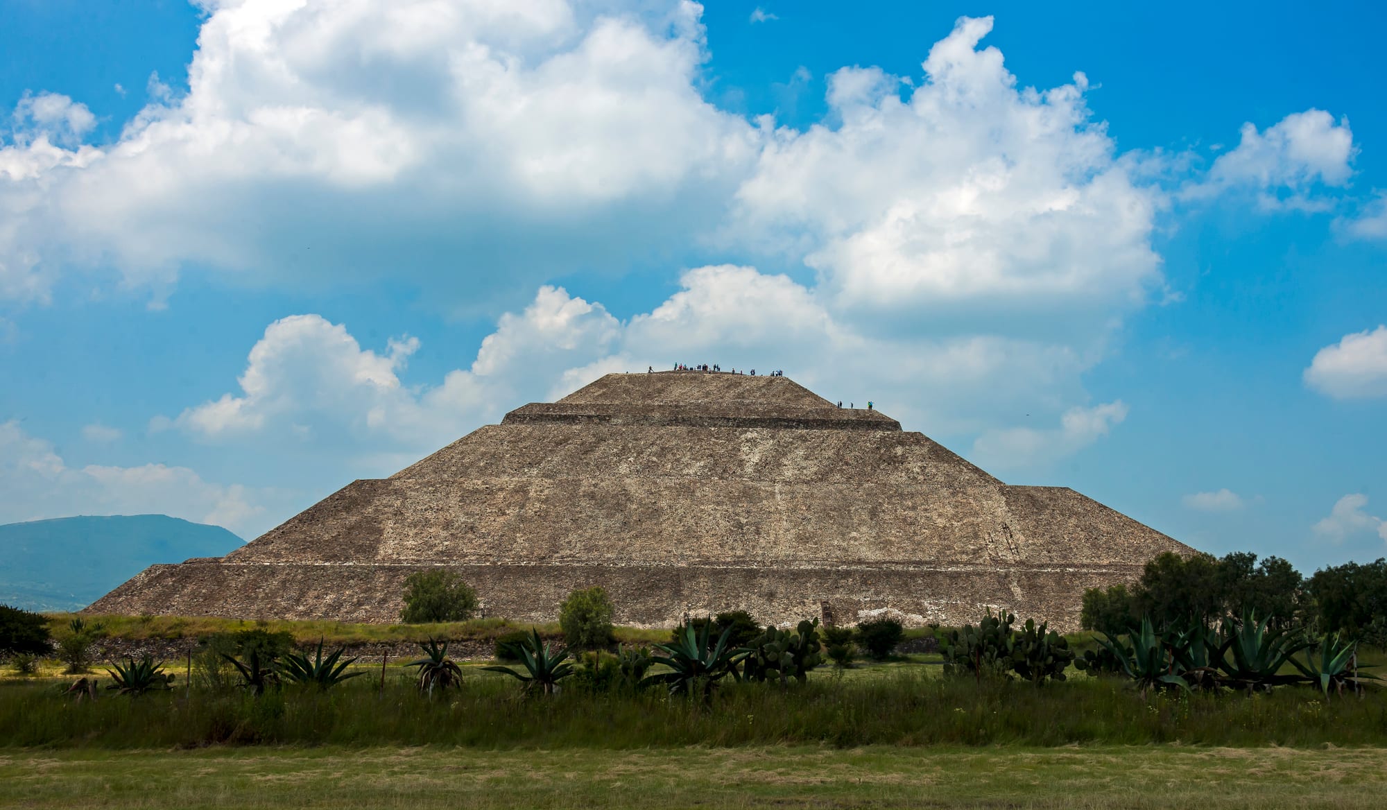 A vast stepped stone pyramid rising from a broad avenue lined with lesser platforms and mountains beyond