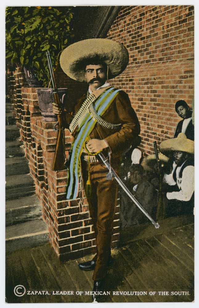 Emiliano Zapata standing with a rifle, sabre, cartridge belts and a wide sombrero