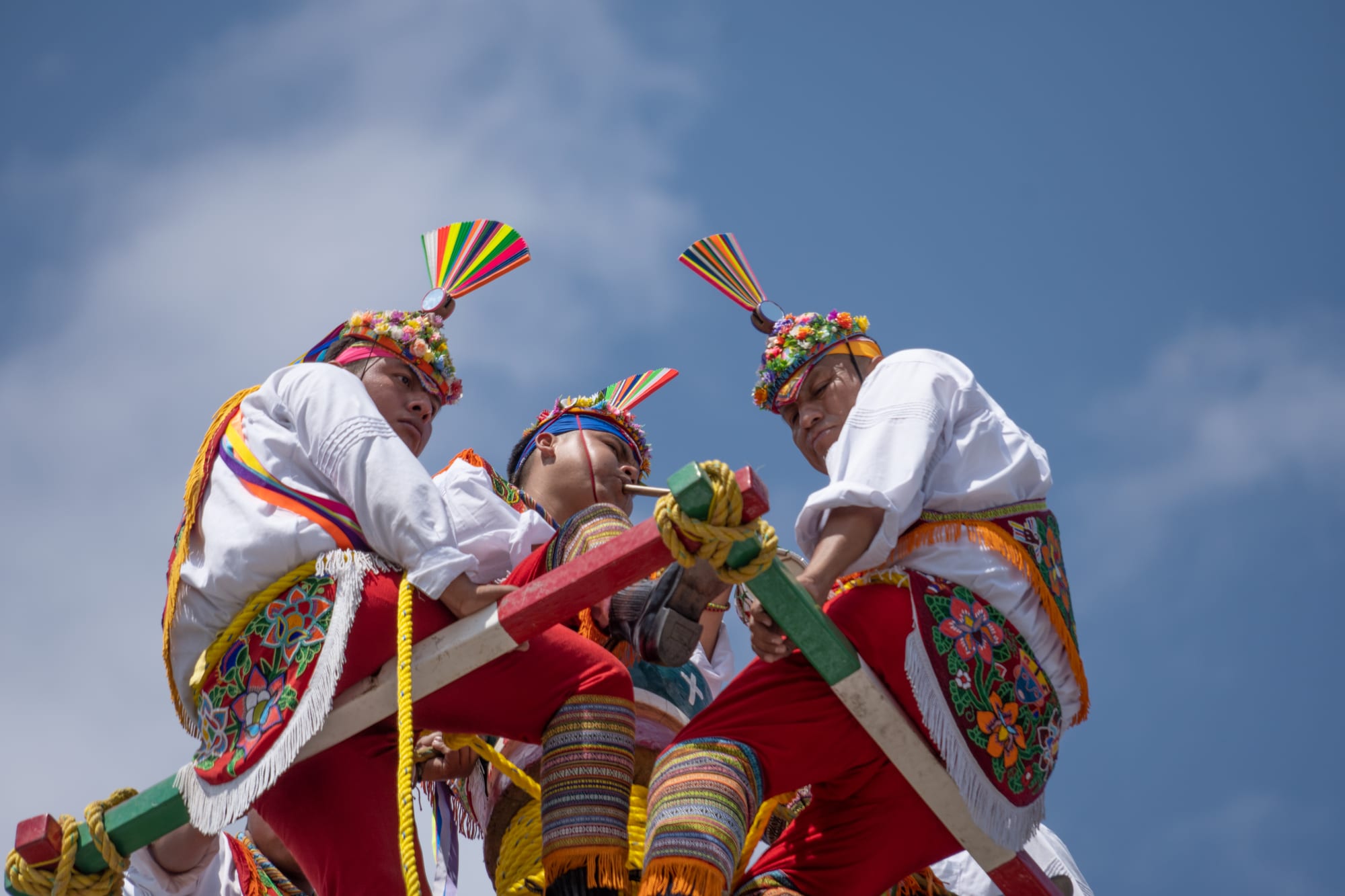 Three men in white shirts, red trousers, embroidered sashes and flower-covered headdresses topped with rainbow-coloured fans sit on a wooden frame at the top of a tall pole, ropes wound around them, against a blue sky