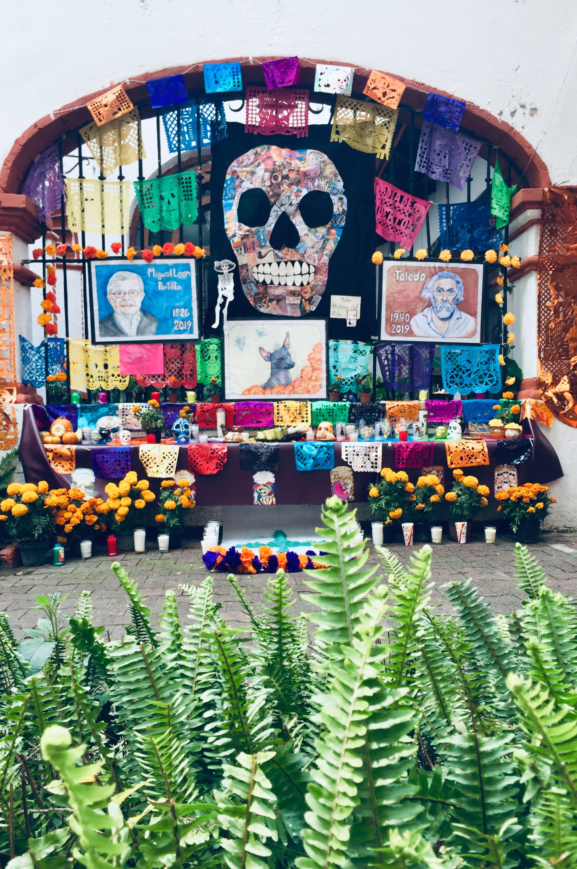 A large Day of the Dead altar under a brick arch, hung with brightly coloured cut-paper banners, with a collage skull, painted portraits, candles and pots of orange marigolds, seen beyond a bank of green ferns