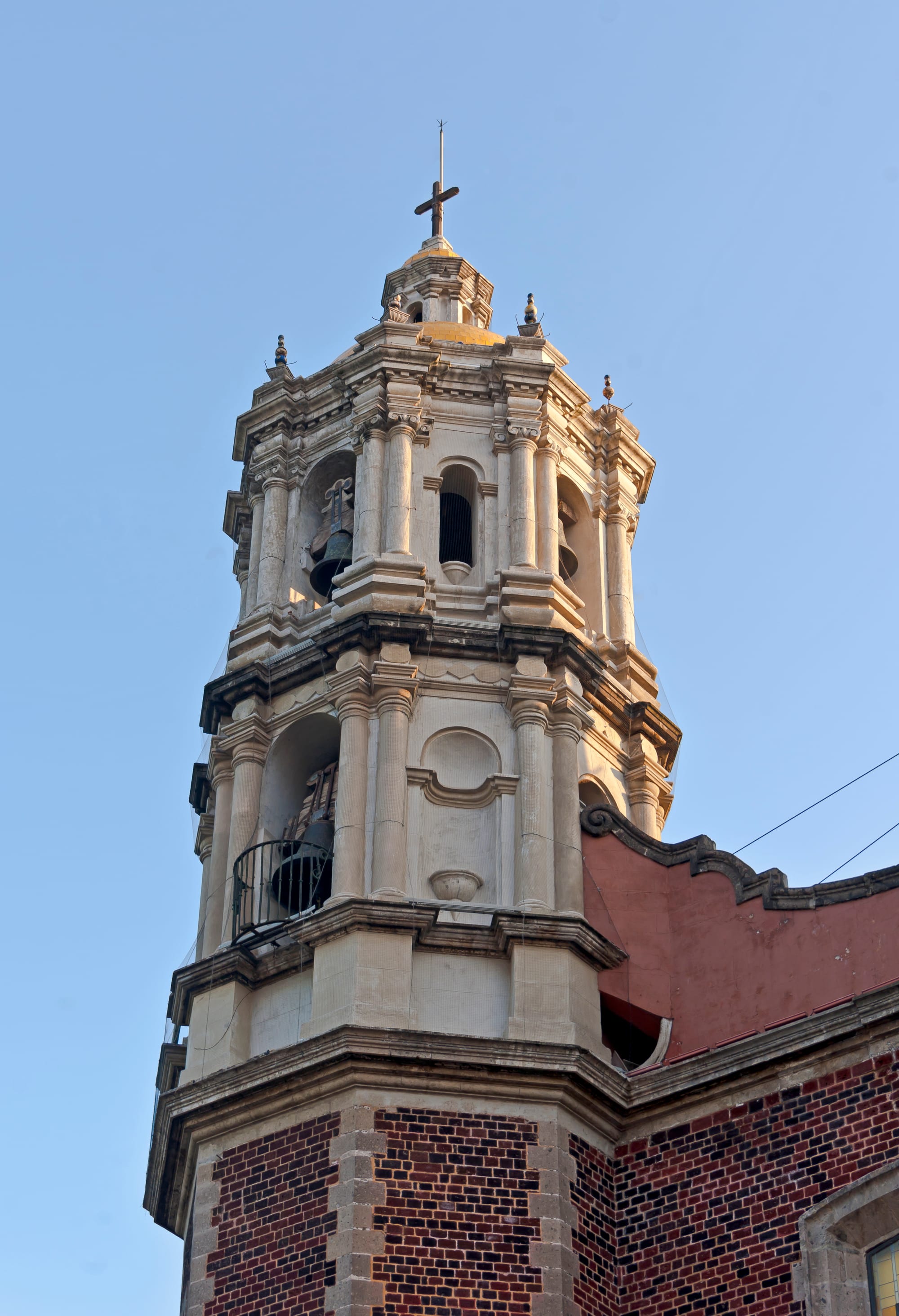 An ornate pale stone baroque bell tower with columns, arched openings holding bells and a cross on top, rising from a building of dark red brick against a clear blue sky