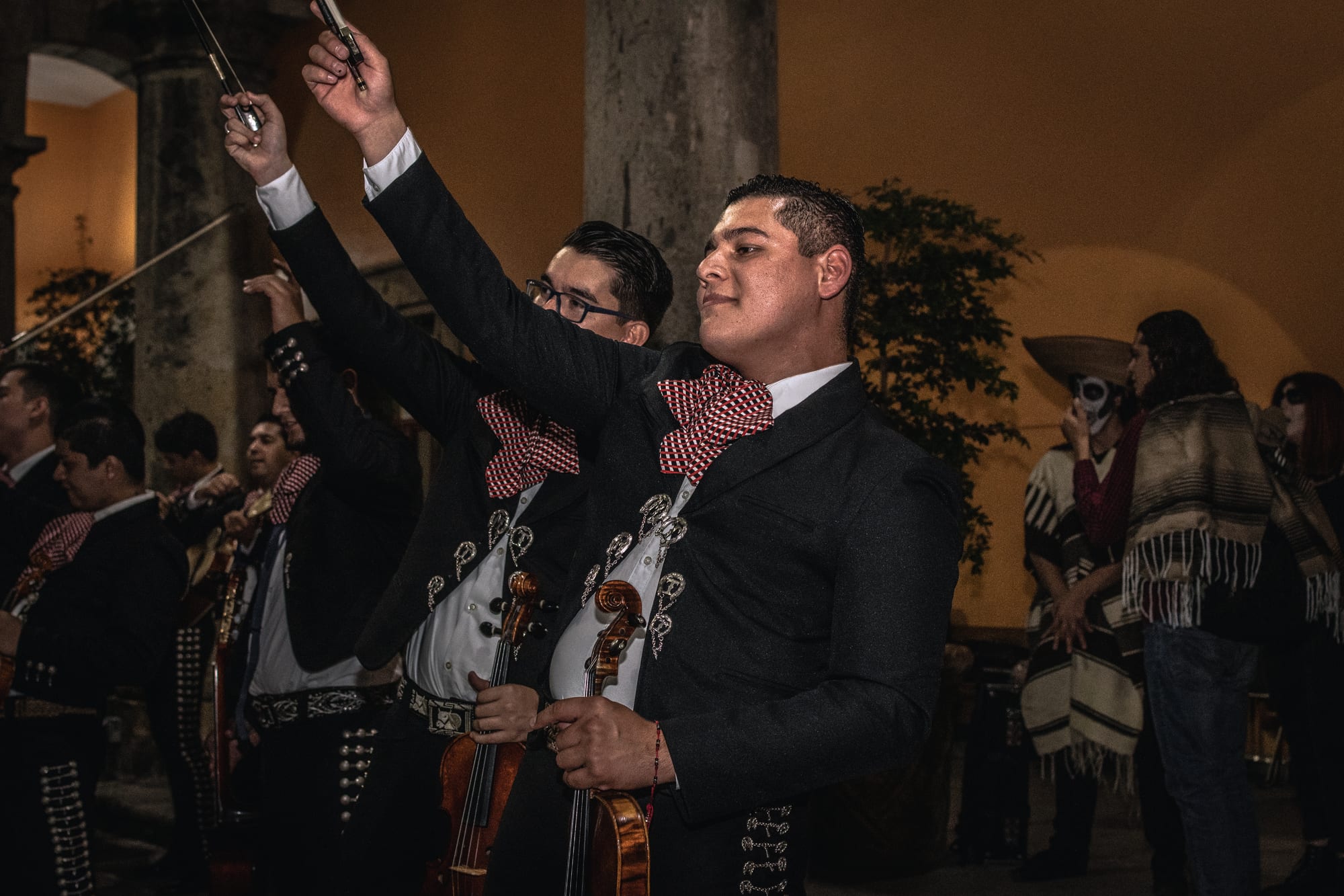 Mariachi musicians in black charro-style suits with silver buttons and red-and-white checked bow ties raise their violin bows high, with other performers and people in skull face paint behind them