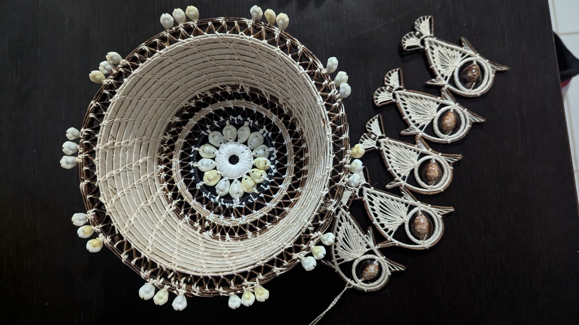 A round woven basket in pale fibre with a dark patterned centre and a rim studded with white shells, next to a string of woven fish-shaped ornaments on a dark table