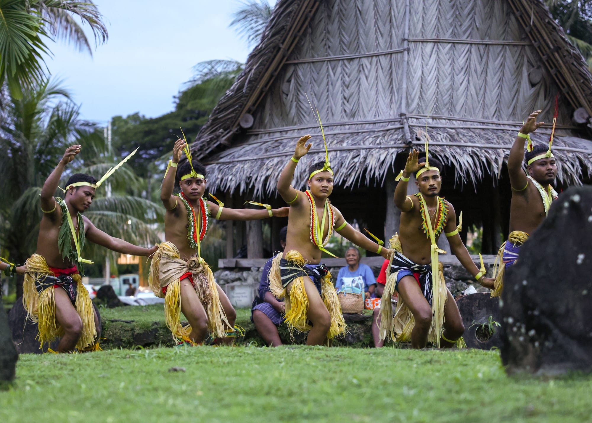 A line of young men in grass skirts, woven necklaces and yellow headbands dancing with one arm raised on a lawn in front of a steep thatched house