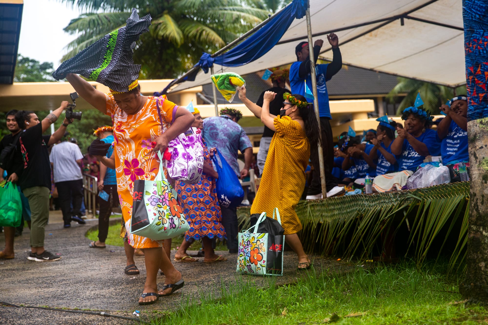 Women in brightly patterned dresses and flower crowns dance and wave colourful cloths on a path beside an open-sided shelter decorated with palm fronds, while others cheer