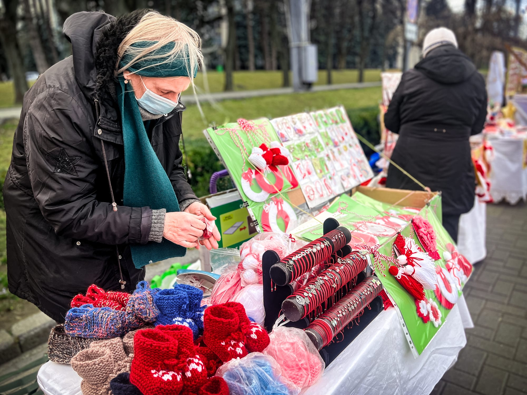 A woman in a dark coat and green scarf arranges small red-and-white thread tokens at a street stall covered with cards of red-and-white charms, knitted red socks and bracelets