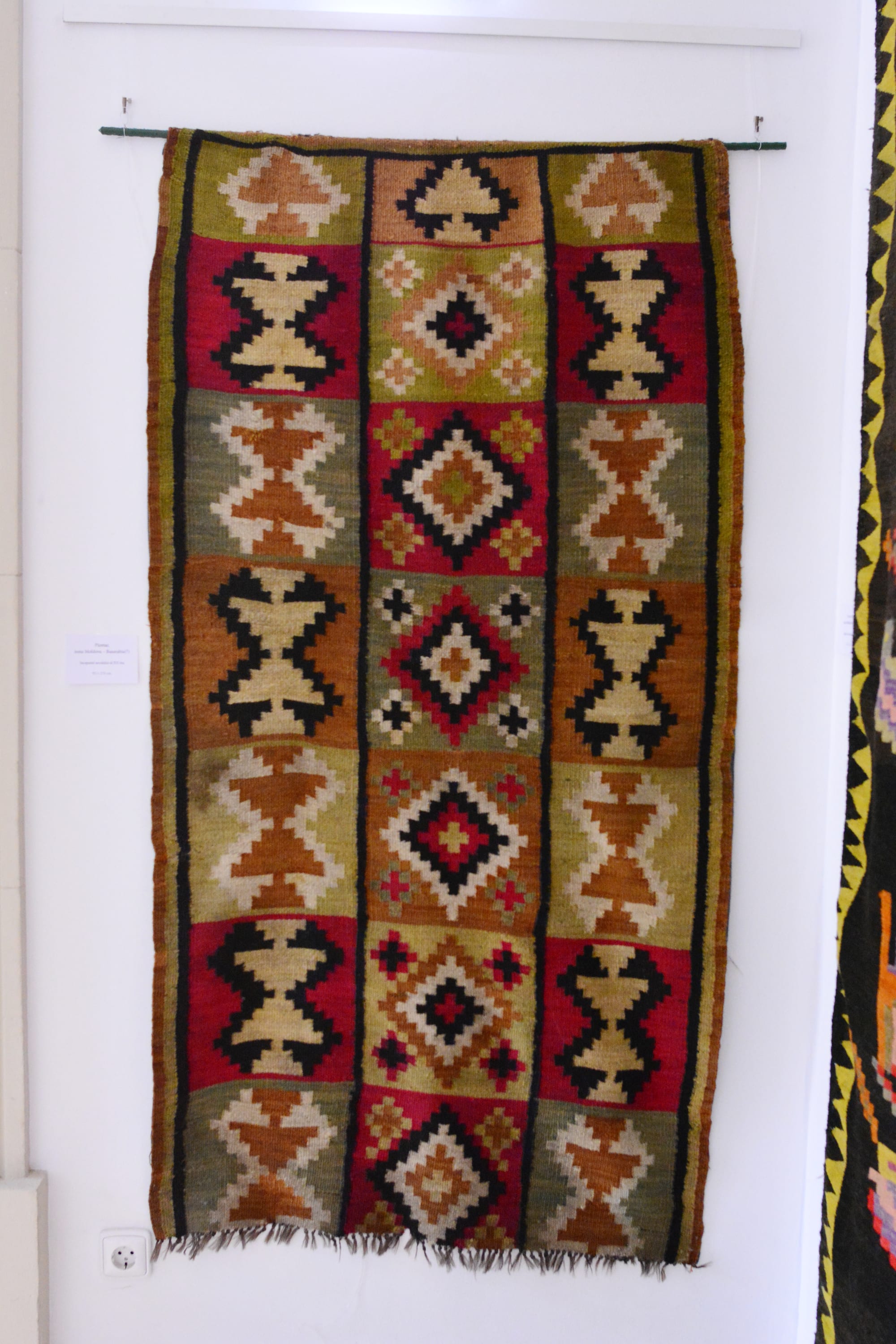 A long woven wall carpet hanging from a rod, patterned in three columns of geometric diamond and hourglass motifs in red, green, ochre, cream and black