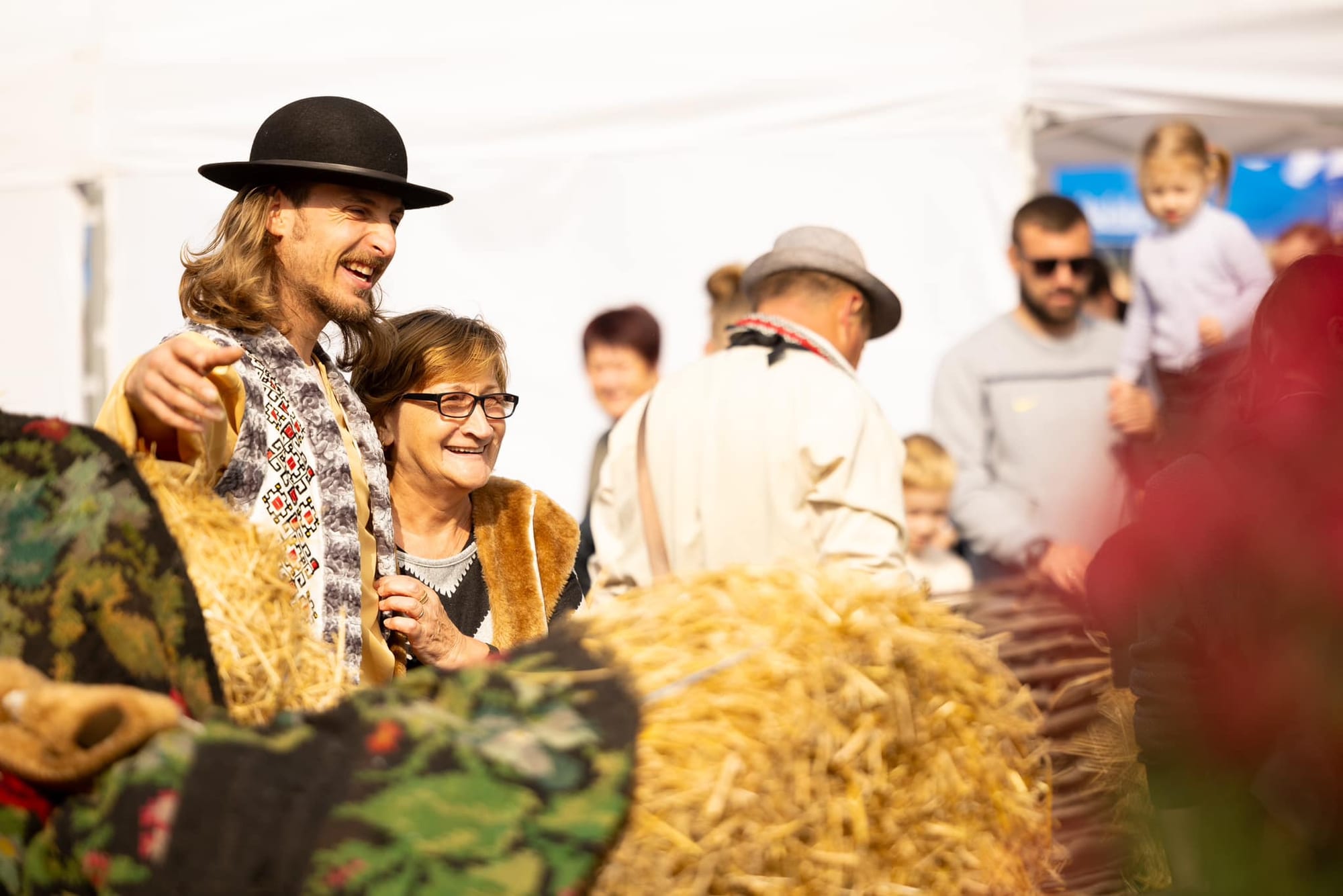 A smiling young man in a black hat and an embroidered white waistcoat puts his arm around an older woman in glasses and a sheepskin waistcoat, beside bales of straw and a flower-patterned rug