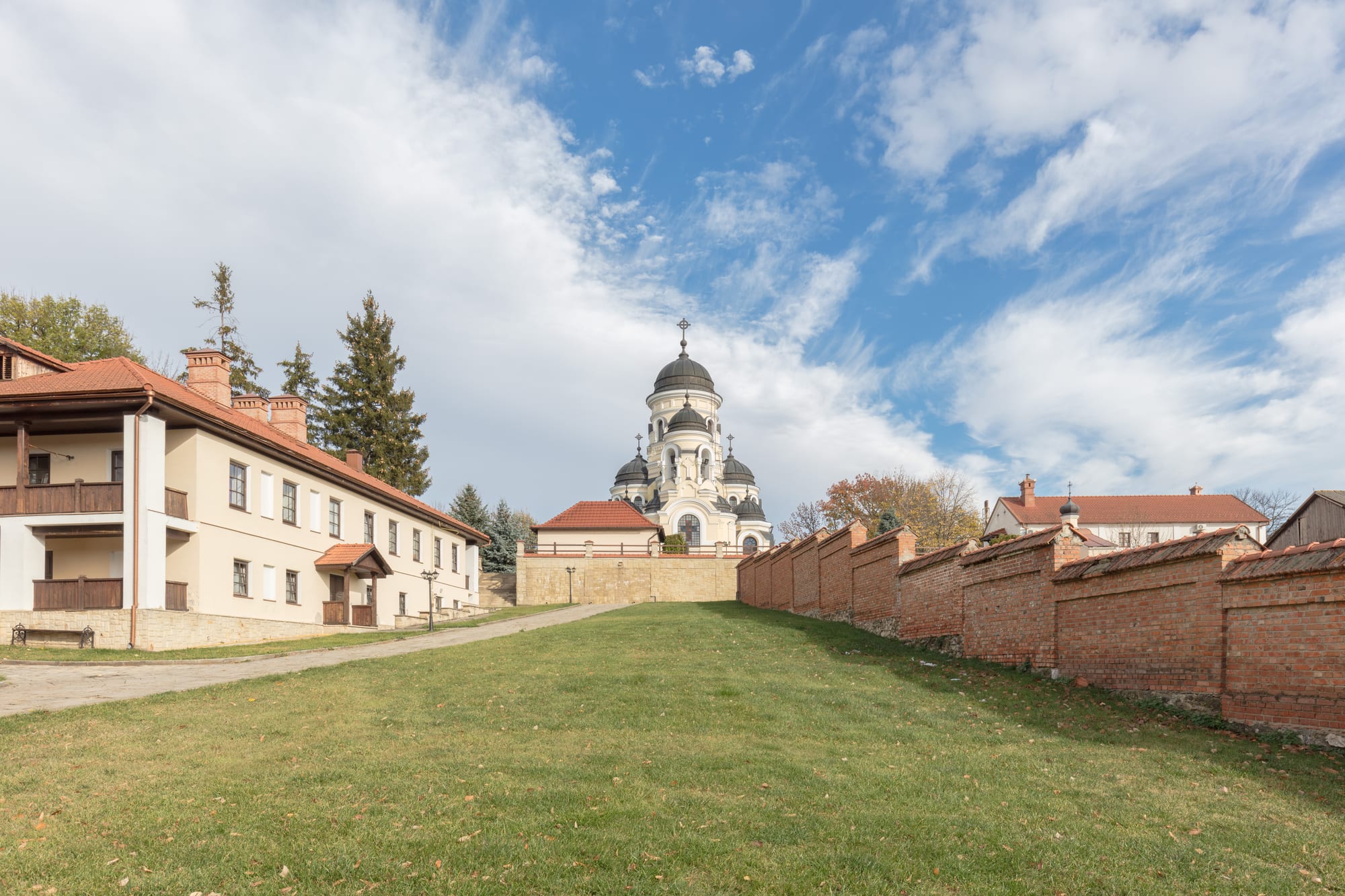 A white church with dark domes and a cross rising beyond a sloping lawn, flanked by a two-storey monastery building on the left and a long red-brick wall on the right under a blue sky