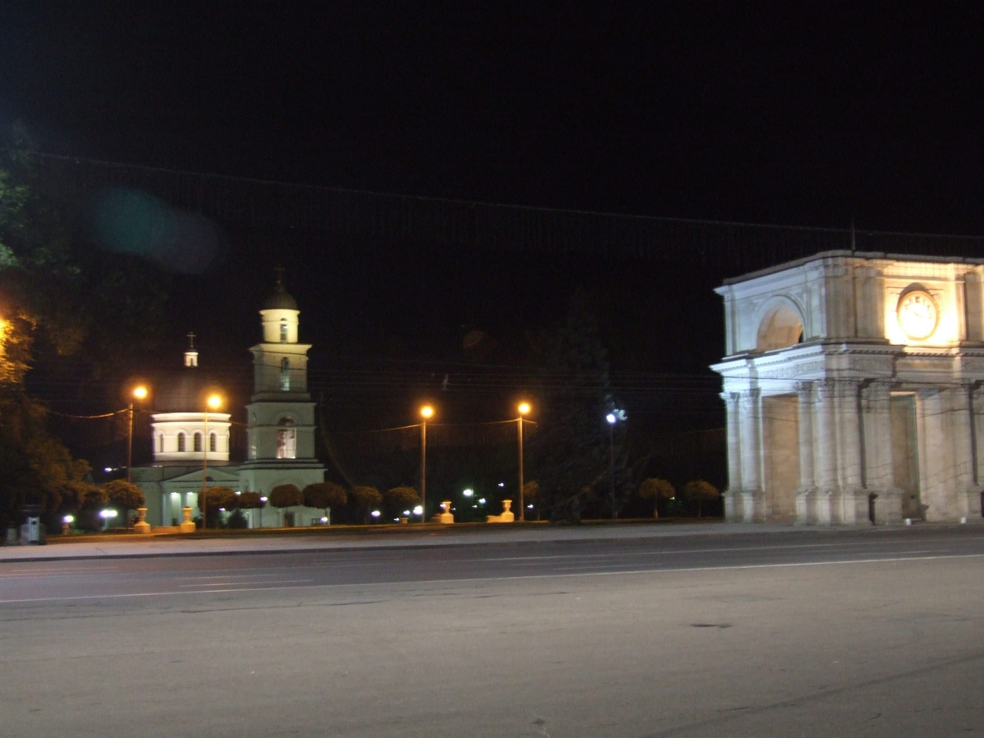A triumphal arch and a cathedral bell tower lit up at night across a square