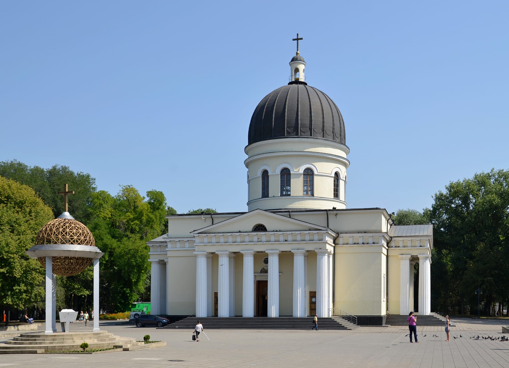 A white neoclassical cathedral with gold domes and a freestanding bell tower on a green square