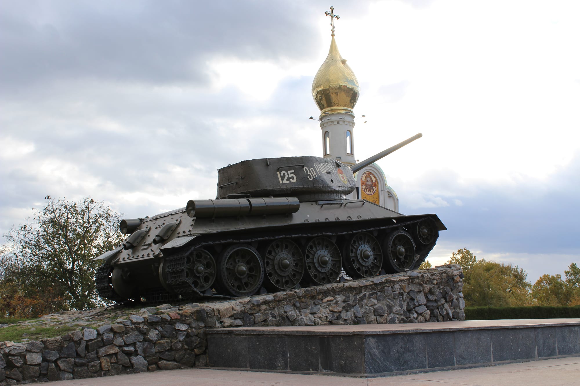 A Soviet-era tank numbered 125 mounted on a stone plinth, with the golden onion dome of a small chapel rising behind it